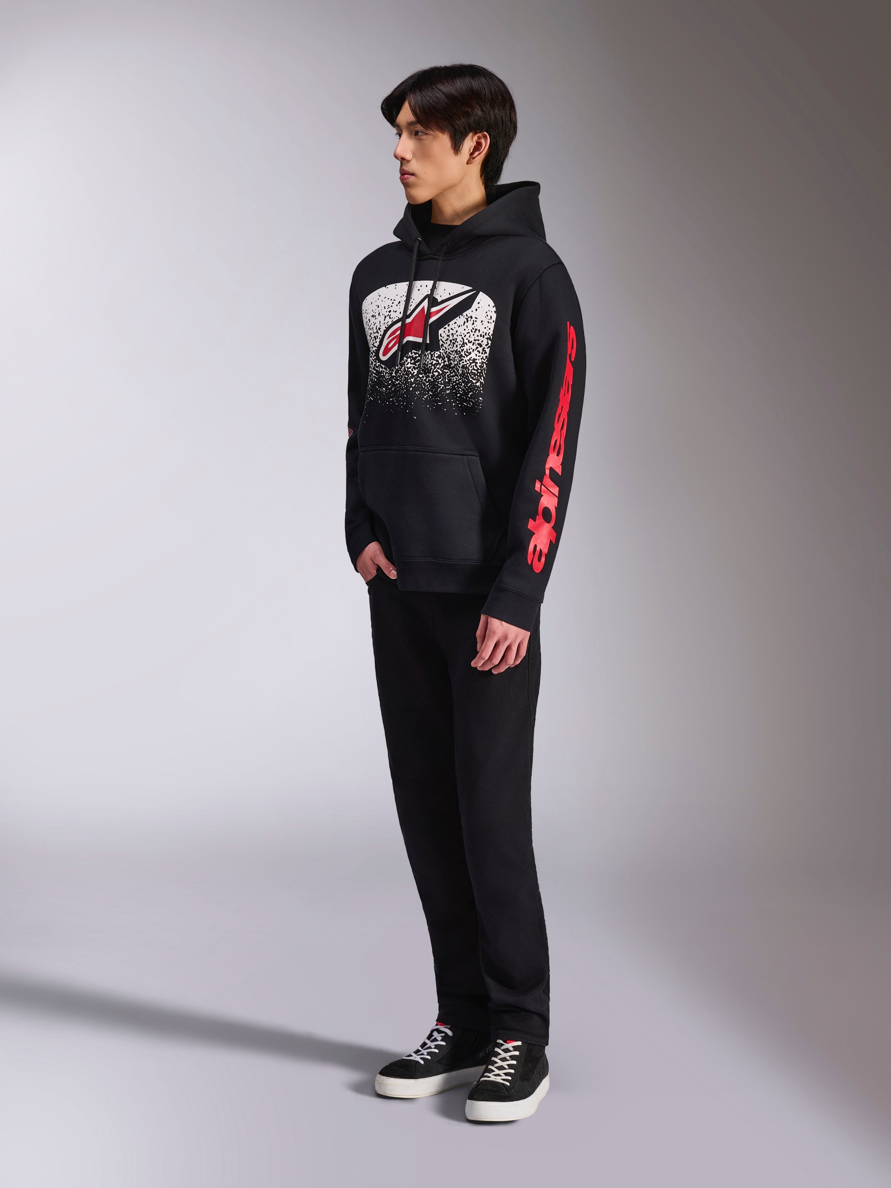 Alpinestars Obsessed Hoodie, zwarte pullover fleece met een grote wit gespikkelde graphic op de borst, rood Astars-logo en rode branding op de mouwen, gedragen door een model met zwarte broek en sneakers, casual motorsport streetwear
