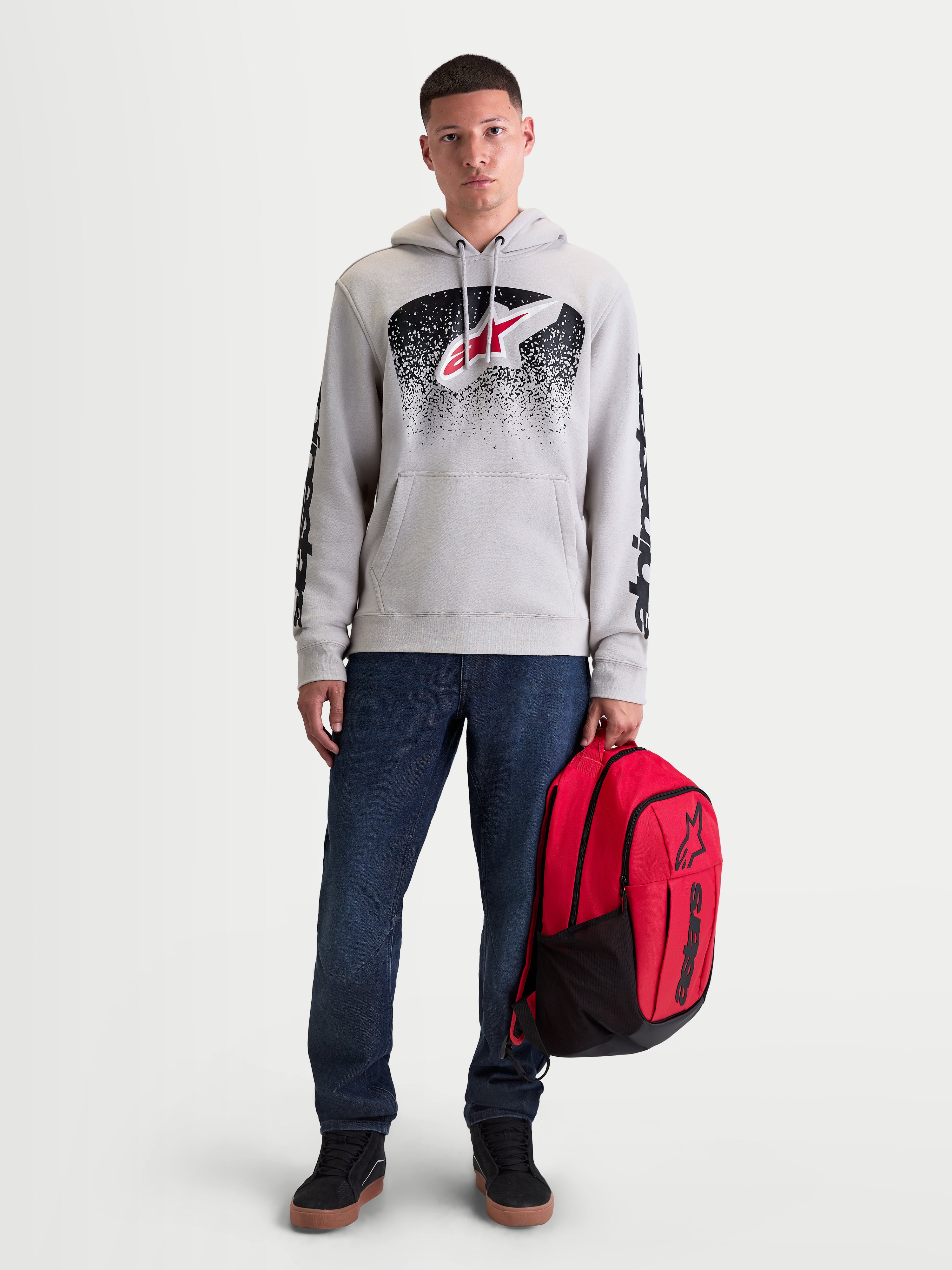 Alpinestars Obsessed hoodie, heren-sportswear pullover fleece, cement grijs met rode en zwarte MX-geïnspireerde verloopgraphics. Deze casual motorsport lifestyle hoodie heeft branding op de mouwen en wordt getoond op een mannelijk model dat een rode Alpinestars-rugzak vasthoudt.