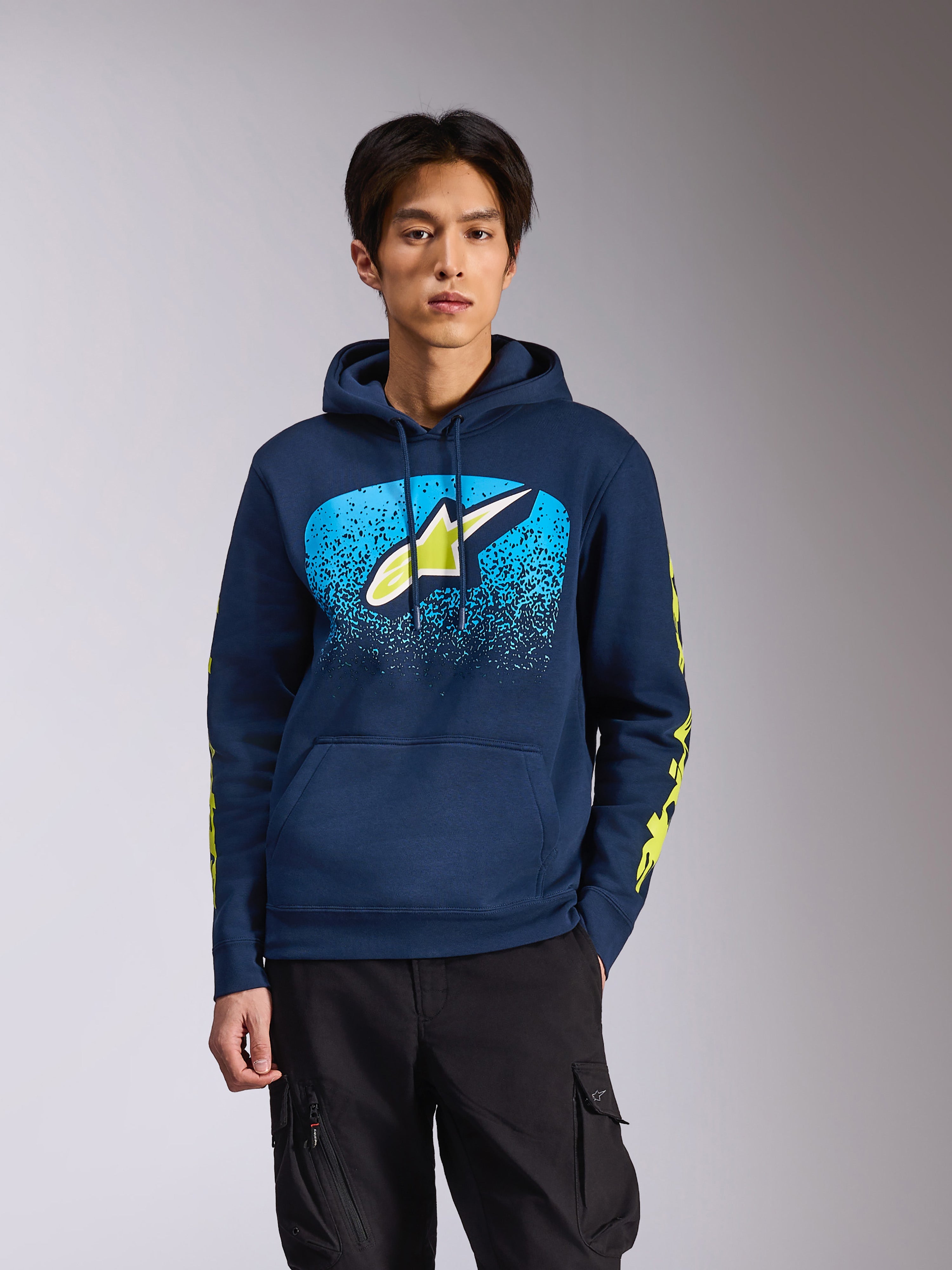 Alpinestars Obsessed Kapuzenpullover in Navy, getragen von einem männlichen Model, mit einem limettengrünen Astars-Logo und hellblauem MX-Farbverlauf auf der Brust sowie gelben Wortmarken-Logos auf den Ärmeln, lässige vom Motorsport inspirierte Streetwear