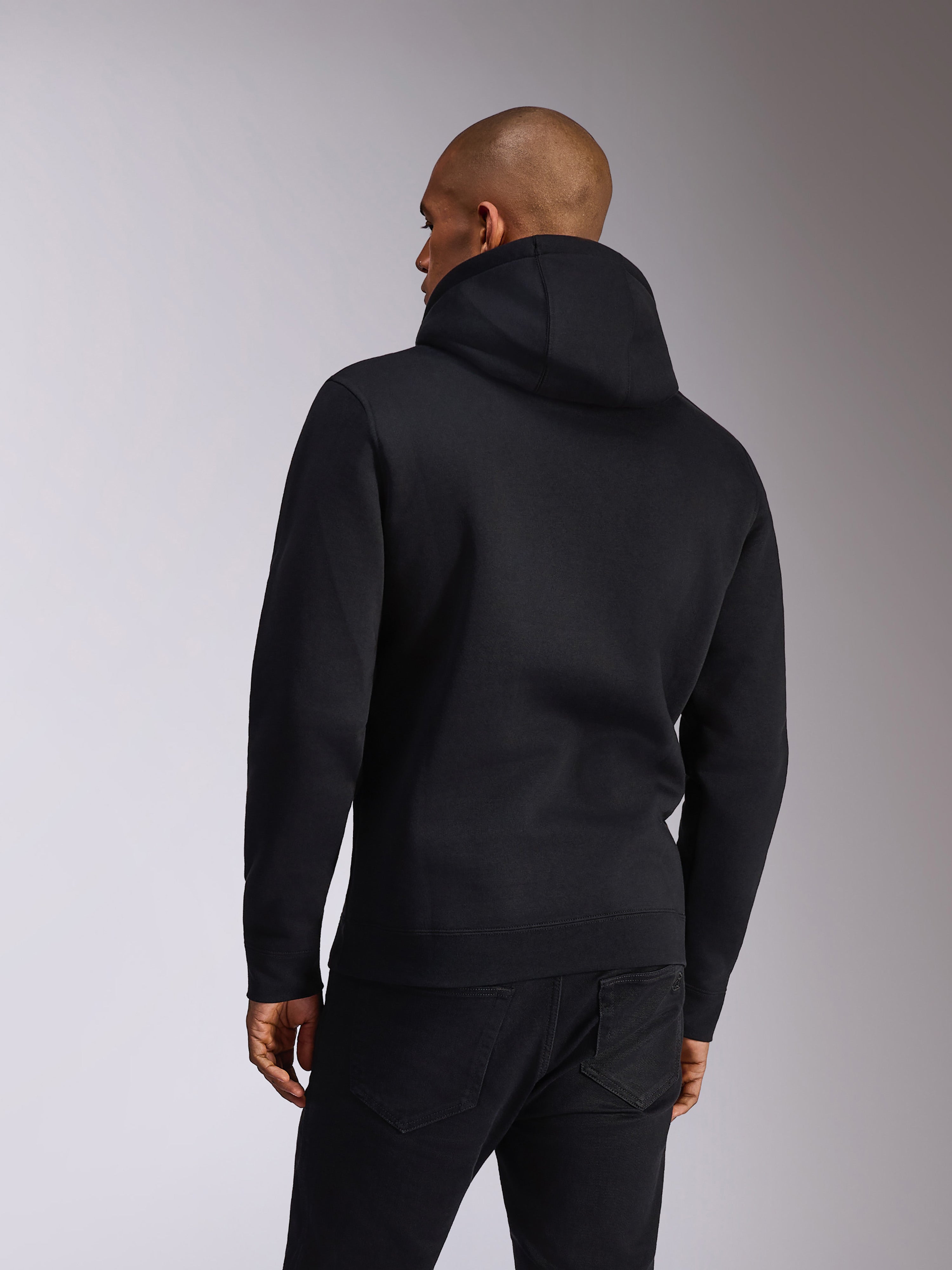 Alpinestars Ellipsoid Hoodie, zwart, achteraanzicht van een persoon die een fleece hoodie draagt, lange mouwen met geribde manchetten en zoom, gecombineerd met een zwarte jeans, casual op de motorsport geïnspireerde sportkleding