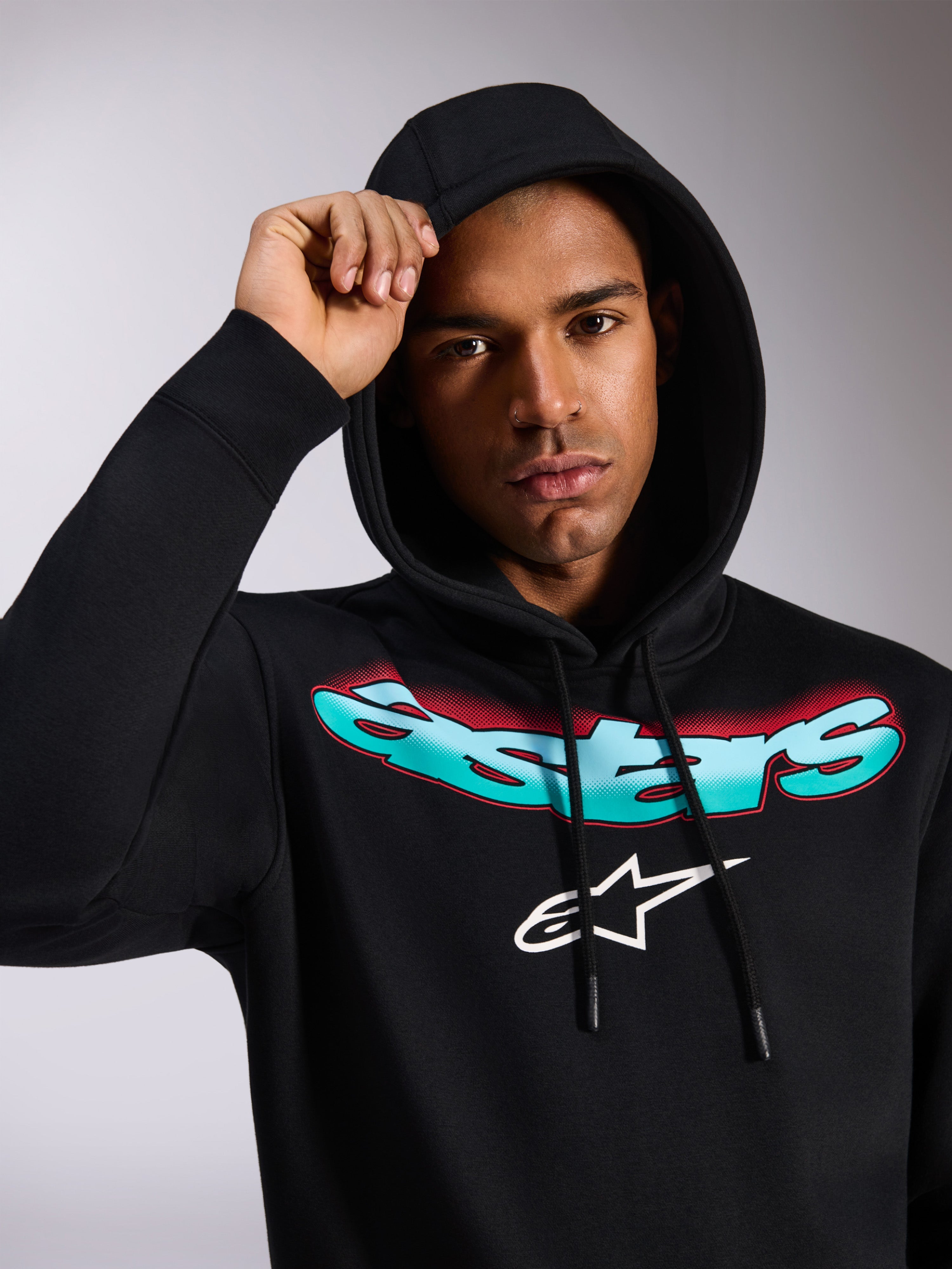 Alpinestars Ellipsoid Hoodie, zwarte pullover fleece hoodie, met gestileerd Astars-woordmerk in blauwgroen en rood kleurverloop en wit logo op de borst, gedragen door model met de capuchon op, casual motorsport-geïnspireerde streetwear