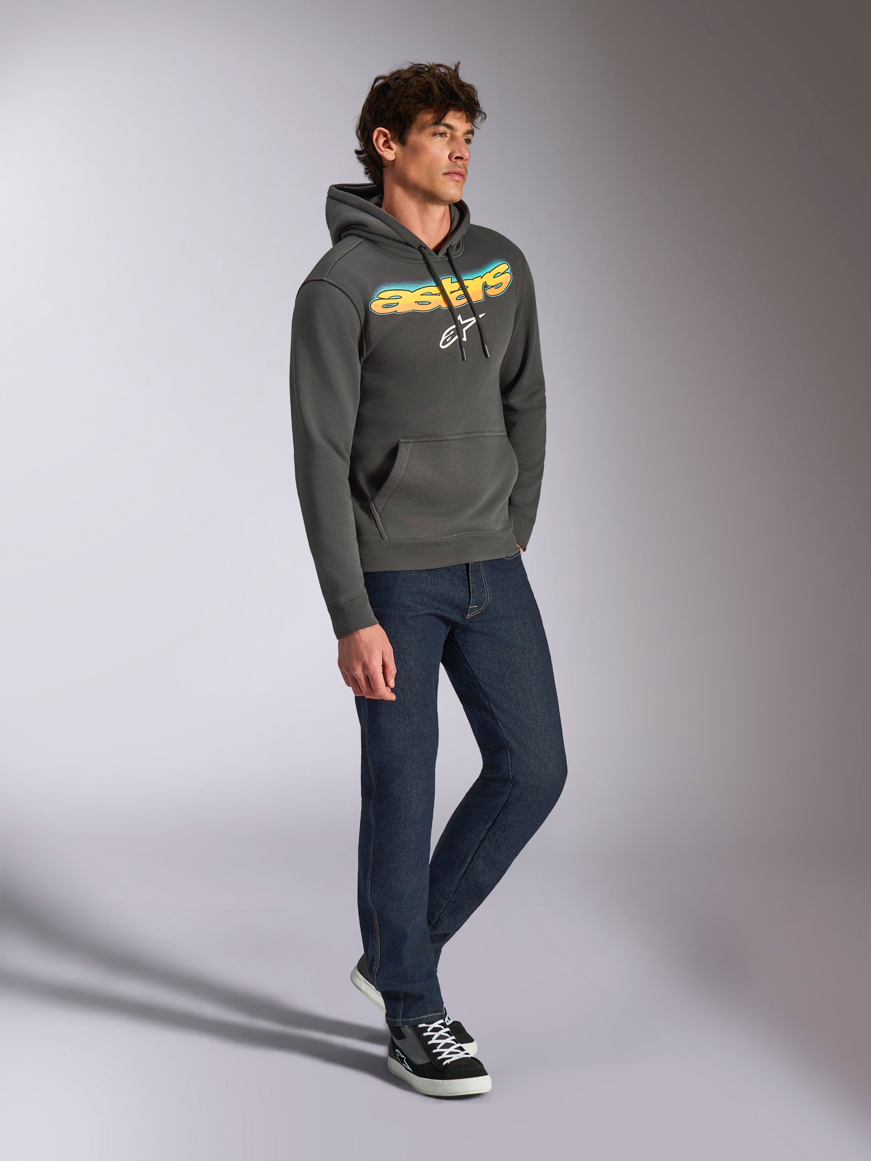 Alpinestars Ellipsoid Hoodie, gunmetal grijze pullover fleece, gebogen Astars-woordmerk aan de voorzijde met een blauw en geel kleurverloop en wit logo, gestyled met een donkere jeans en zwarte sneakers, casual streetwear.