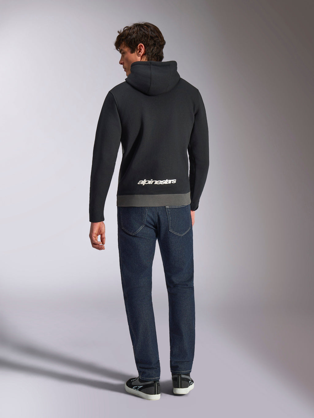 Alpinestars Parallel Hoodie, achteraanzicht van een man in een zwart en gunmetal kleurgeblokte fleece trui, wit Alpinestars woordlogo op de onderrug, gecombineerd met een donkerblauwe jeans, casual op motorsport geïnspireerde streetwear