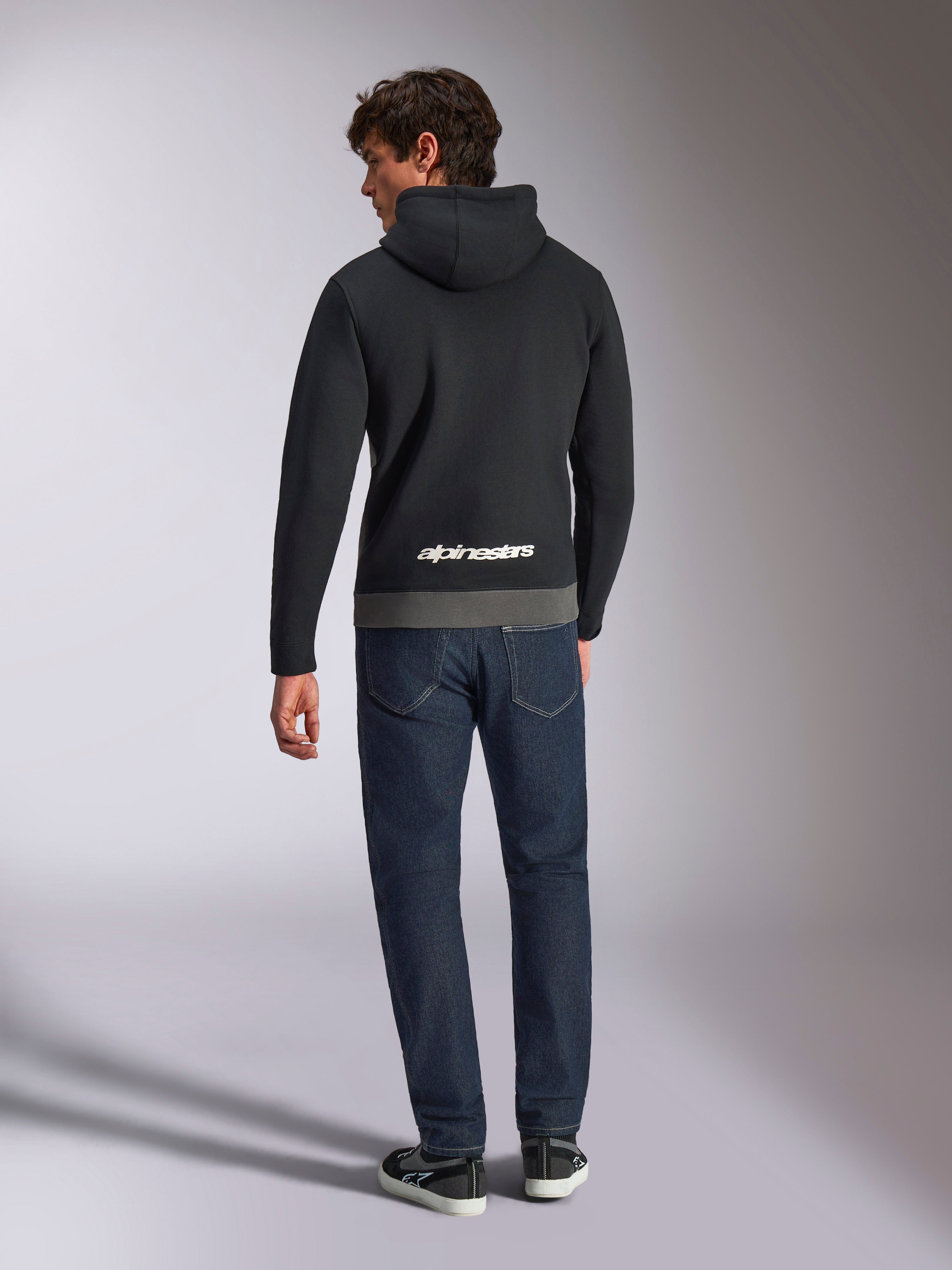 Alpinestars Parallel Hoodie, achteraanzicht van een man in een zwart en gunmetal kleurgeblokte fleece trui, wit Alpinestars woordlogo op de onderrug, gecombineerd met een donkerblauwe jeans, casual op motorsport geïnspireerde streetwear