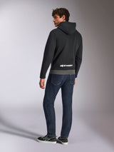 Alpinestars Parallel Hoodie, achteraanzicht van model in de zwart en gunmetal grijze pullover, voorzien van wit Alpinestars-logo op de onderrug, gecombineerd met donkerblauwe jeans, casual motorsport-geïnspireerde streetwear