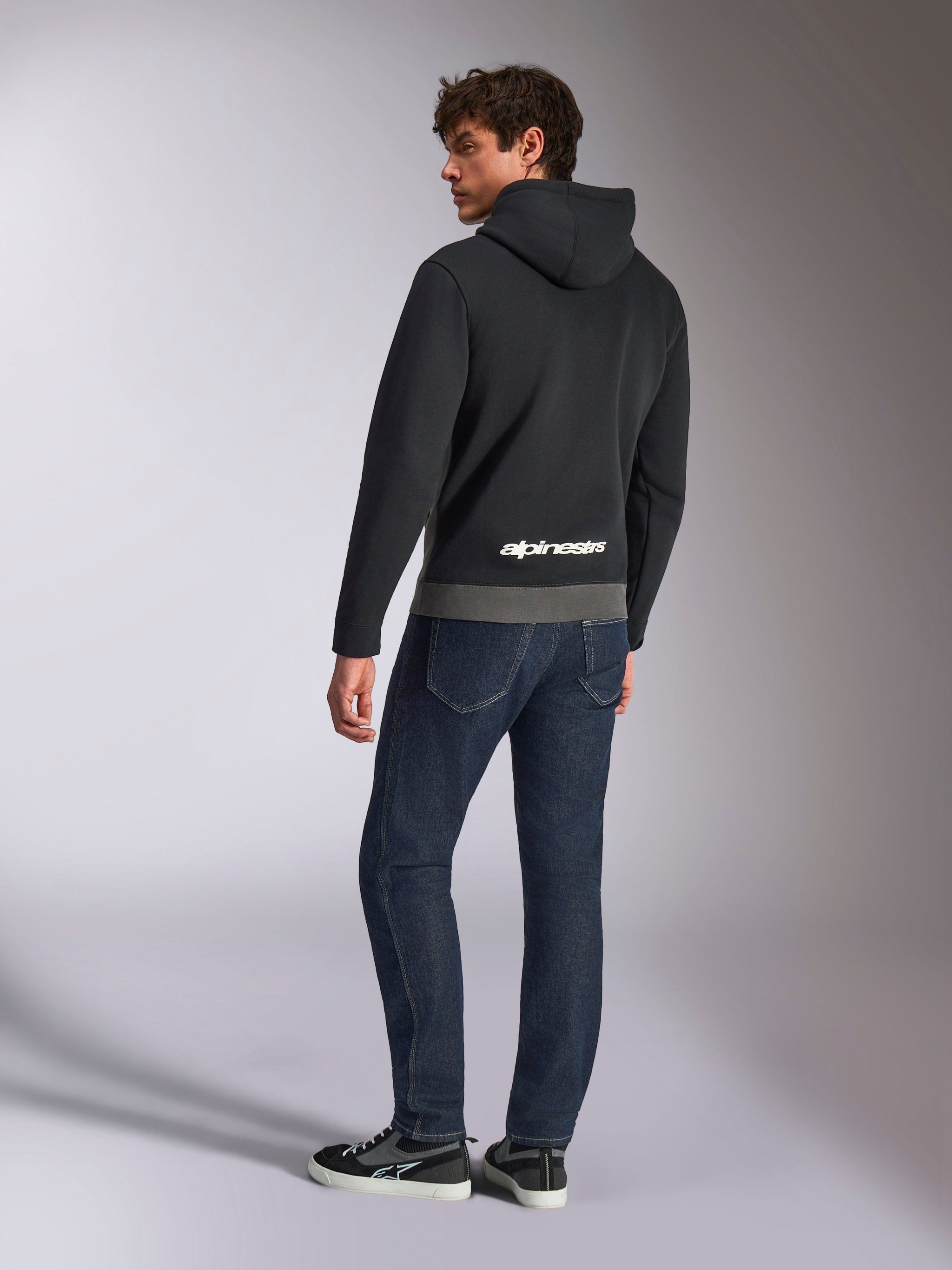 Alpinestars Parallel Hoodie, achteraanzicht van model in de zwart en gunmetal grijze pullover, voorzien van wit Alpinestars-logo op de onderrug, gecombineerd met donkerblauwe jeans, casual motorsport-geïnspireerde streetwear