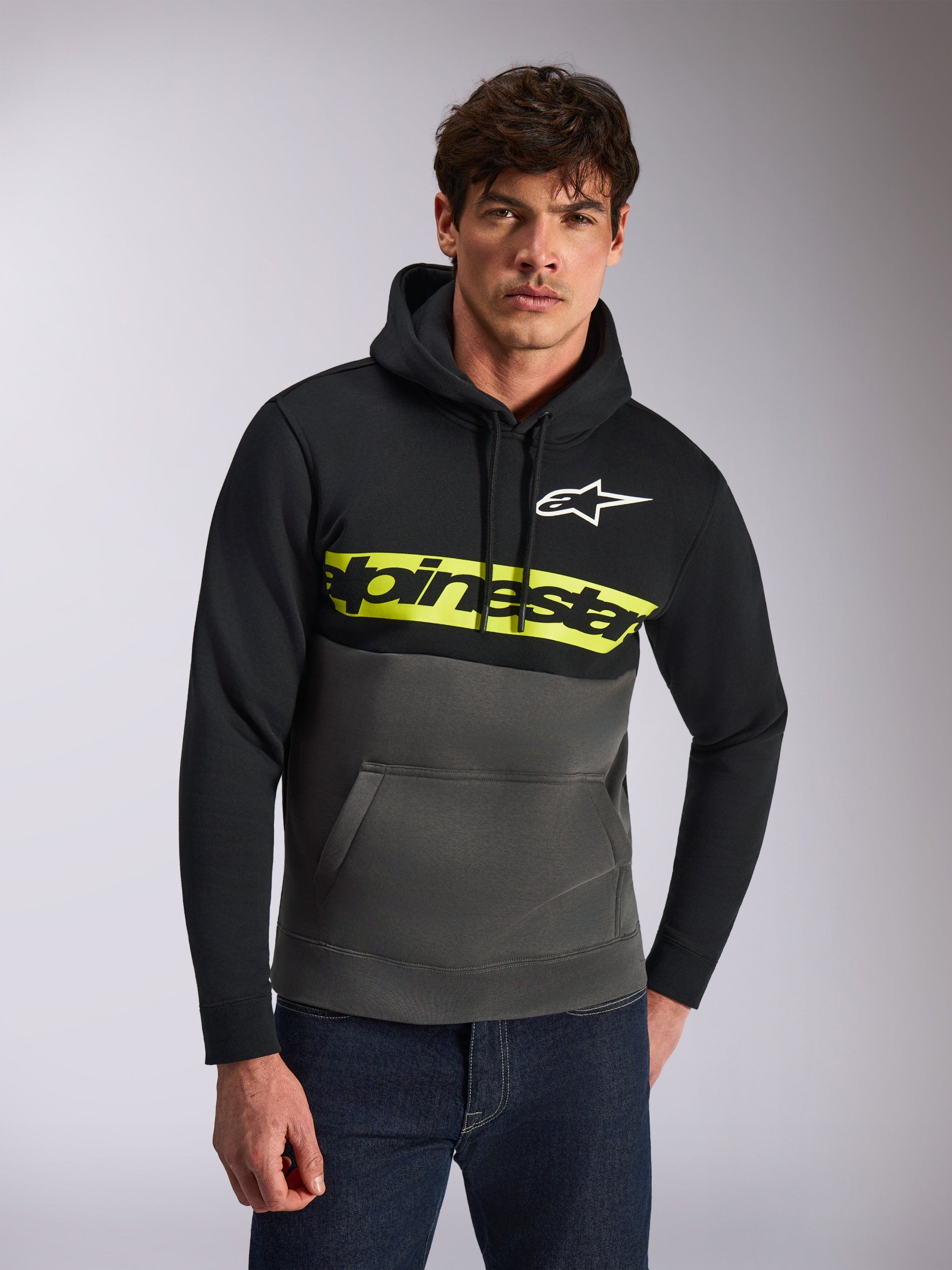 Alpinestars Parallel Hoodie, casual streetwear, kleurgeblokkeerd zwart en gunmetal grijs, felgele borststreep met Alpinestars-woordlogo, wit Astars-logo, gedragen door een model met blauwe jeans, modern op motorsport geïnspireerd ontwerp