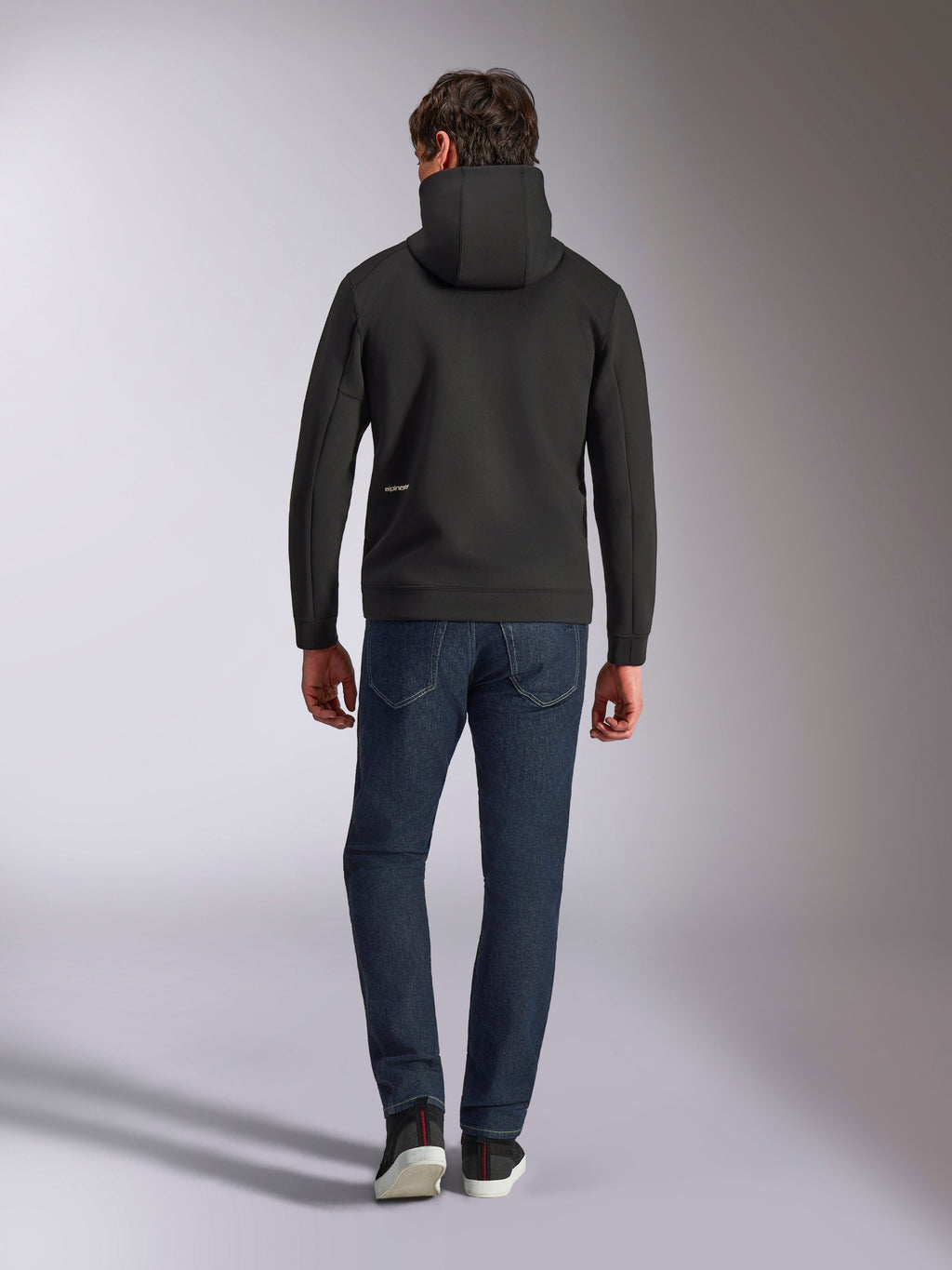 Alpinestars Cypher Hoodie, zwarte fleece met dubbele stof, achteraanzicht met een klein wit Alpinestars-logo op de linkerheup, hoge capuchon, gecombineerd met een donkerblauwe jeans, casual streetwear geïnspireerd op de motorsport