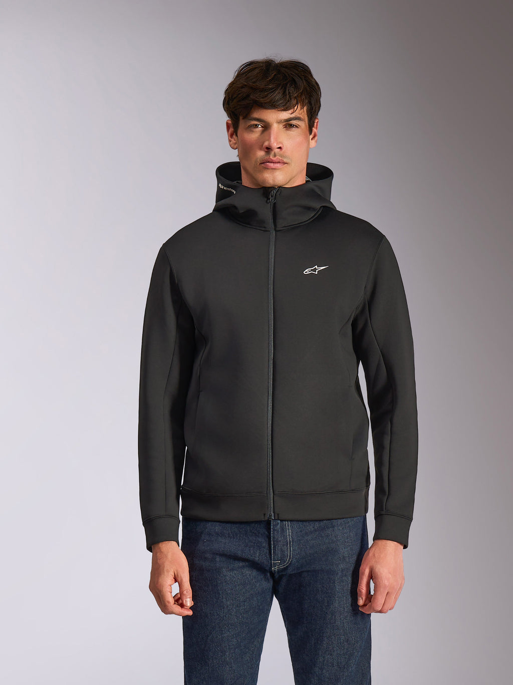 Alpinestars Cypher Hoodie, zwarte fleece hoodie met ritssluiting aan de voorzijde, voorzien van een hoge capuchon met witte branding en een klein wit Astars-logo op de borst, gedragen door een model met blauwe jeans, casual op motorsport geïnspireerde streetwear.