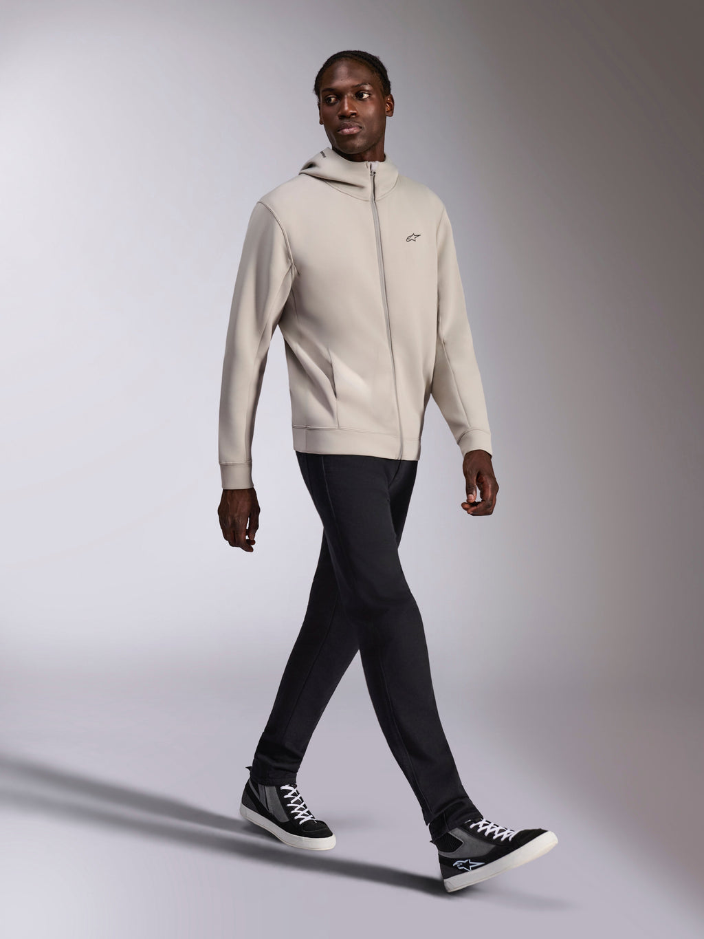 Alpinestars Cypher Hoodie, Cement grijs fleecevest met rits, hoge capuchon, klein Astars-logo op de borst, gedragen door een lopend model, gecombineerd met een zwarte broek en zwart-grijze sneakers, casual motorsport-geïnspireerde sportkleding.