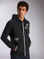 Alpinestars Transverse Hoodie, casual sportkleding, zwart met wit ritsdetail, capuchon met trekkoord, Alpinestars ster-logo op de borst, 'One Goal One Vision' graphic, gedragen door een mannelijk model met donker krullend haar