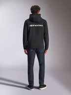 Alpinestars Transverse Hoodie, casual motorsport streetwear, zwart met wit Alpinestars-woordlogo op de rug, lange mouwen, geribde boorden en manchetten, van achteren gedragen met een donkere denim jeans