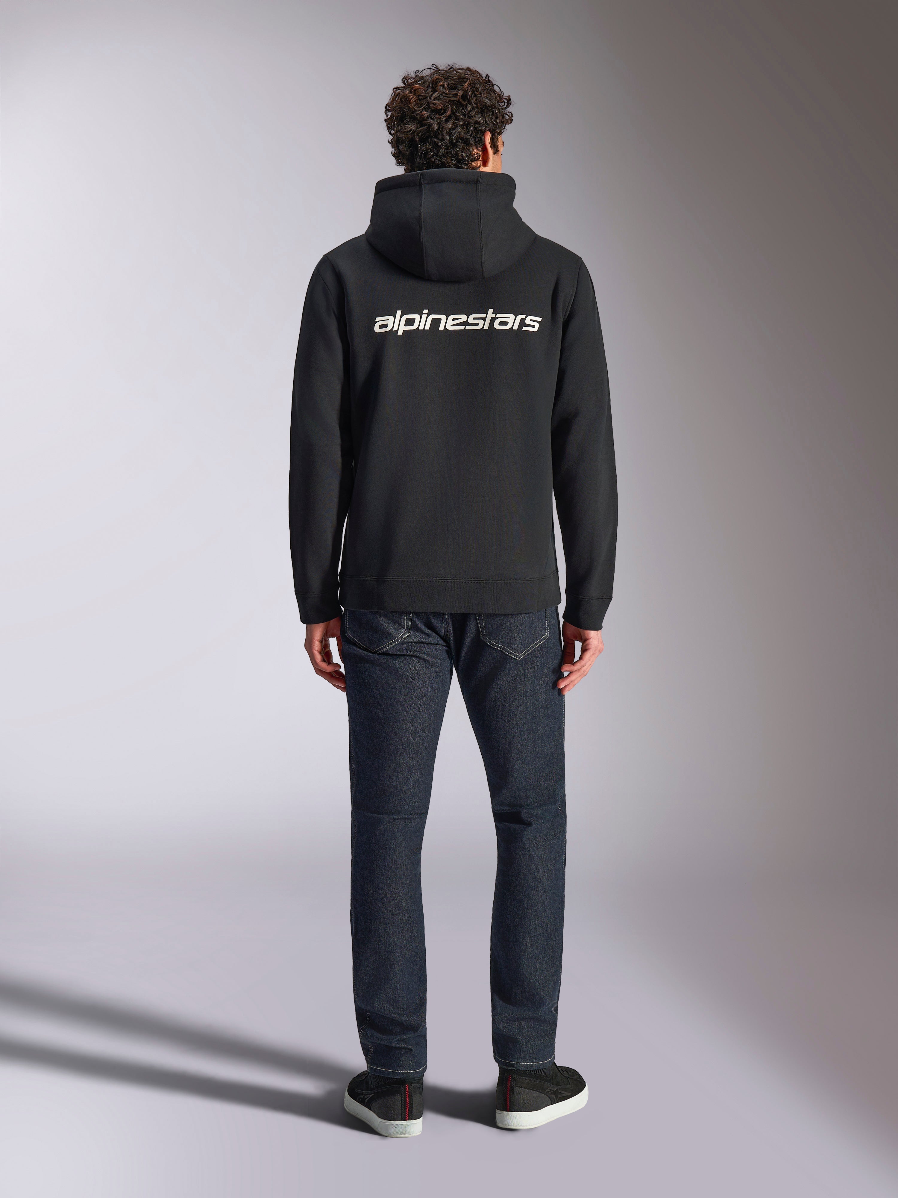 Alpinestars Transverse Hoodie, casual motorsport streetwear, zwart met wit Alpinestars-woordlogo op de rug, lange mouwen, geribde boorden en manchetten, van achteren gedragen met een donkere denim jeans