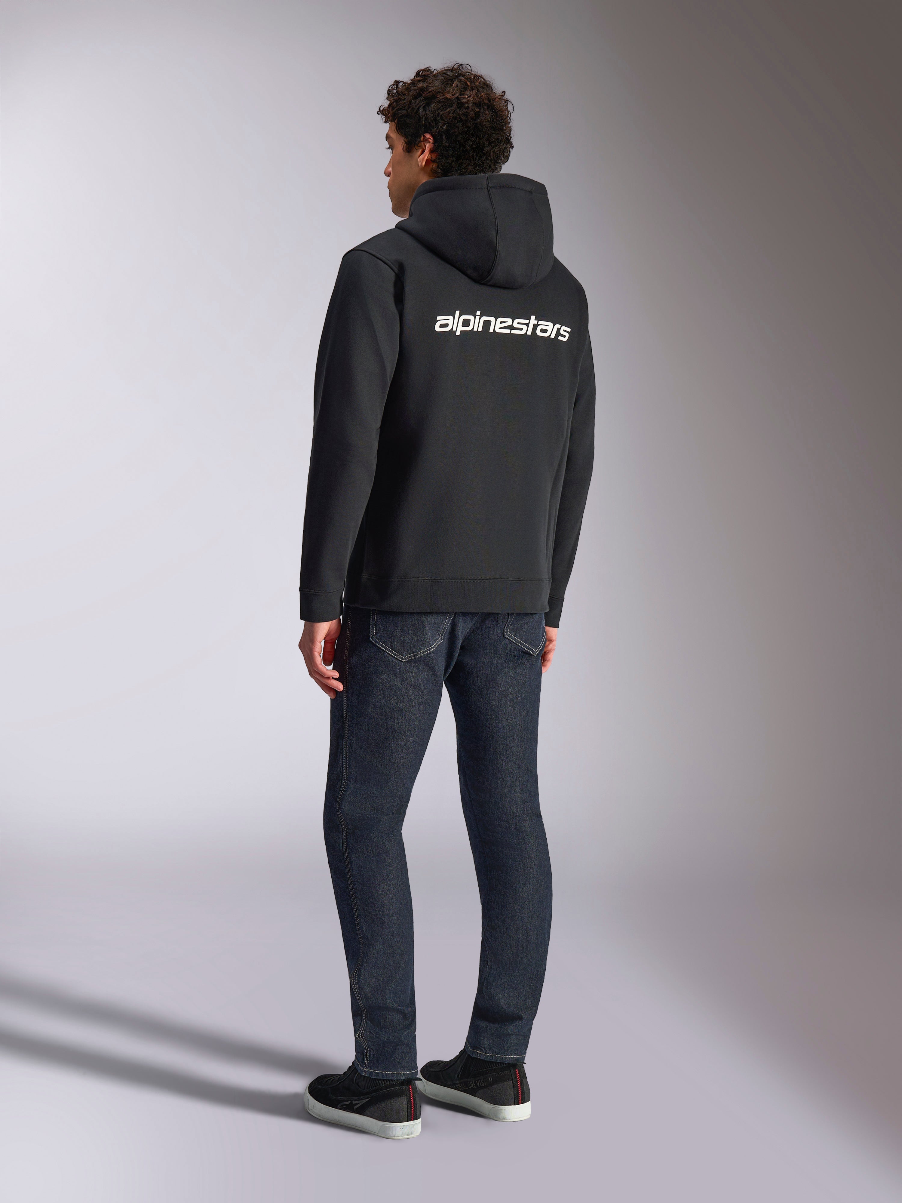 Alpinestars Transverse Hoodie, zwarte fleece hoodie met rits, achteraanzicht met een wit Alpinestars-woordlogo over de schouders, gedragen met donkerblauwe jeans en zwarte sneakers, casual op motorsport geïnspireerde streetwear