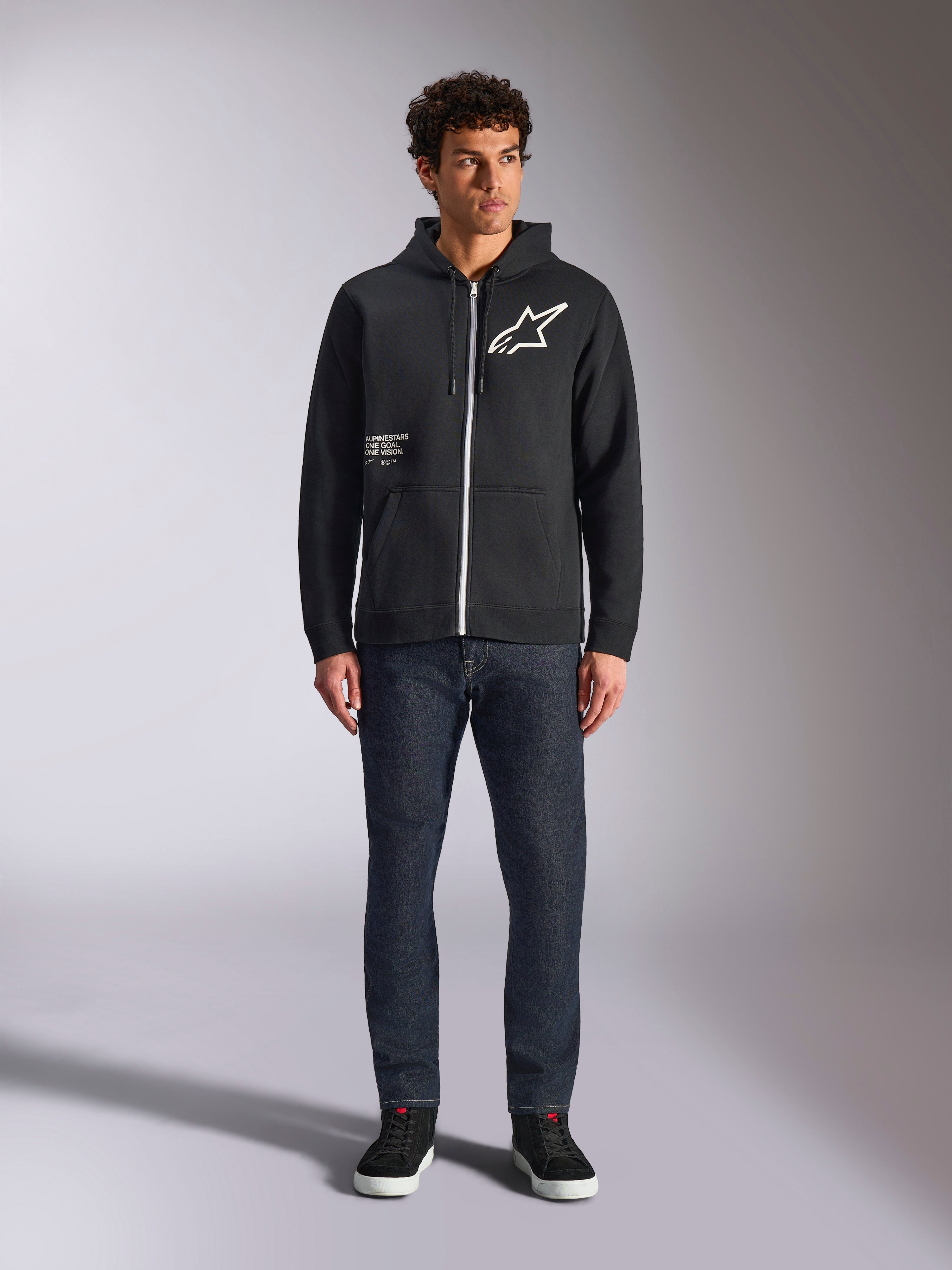 Alpinestars Transverse Hoodie, casual fleece hoodie met rits, zwart, wit Astars-logo op de borst en 'One Goal'-print aan de voorkant, capuchon met trekkoord, gecombineerd met een donkere jeans en zwarte sneakers, lifestyle motorsport streetwear