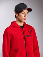 Sweat à capuche Alpinestars Transverse, polaire rouge à fermeture éclair frontale, logo Astars noir sur la poitrine et texte 'One Goal. One Vision.' sur le bas avant, style streetwear décontracté, porté par un mannequin avec une casquette noire
