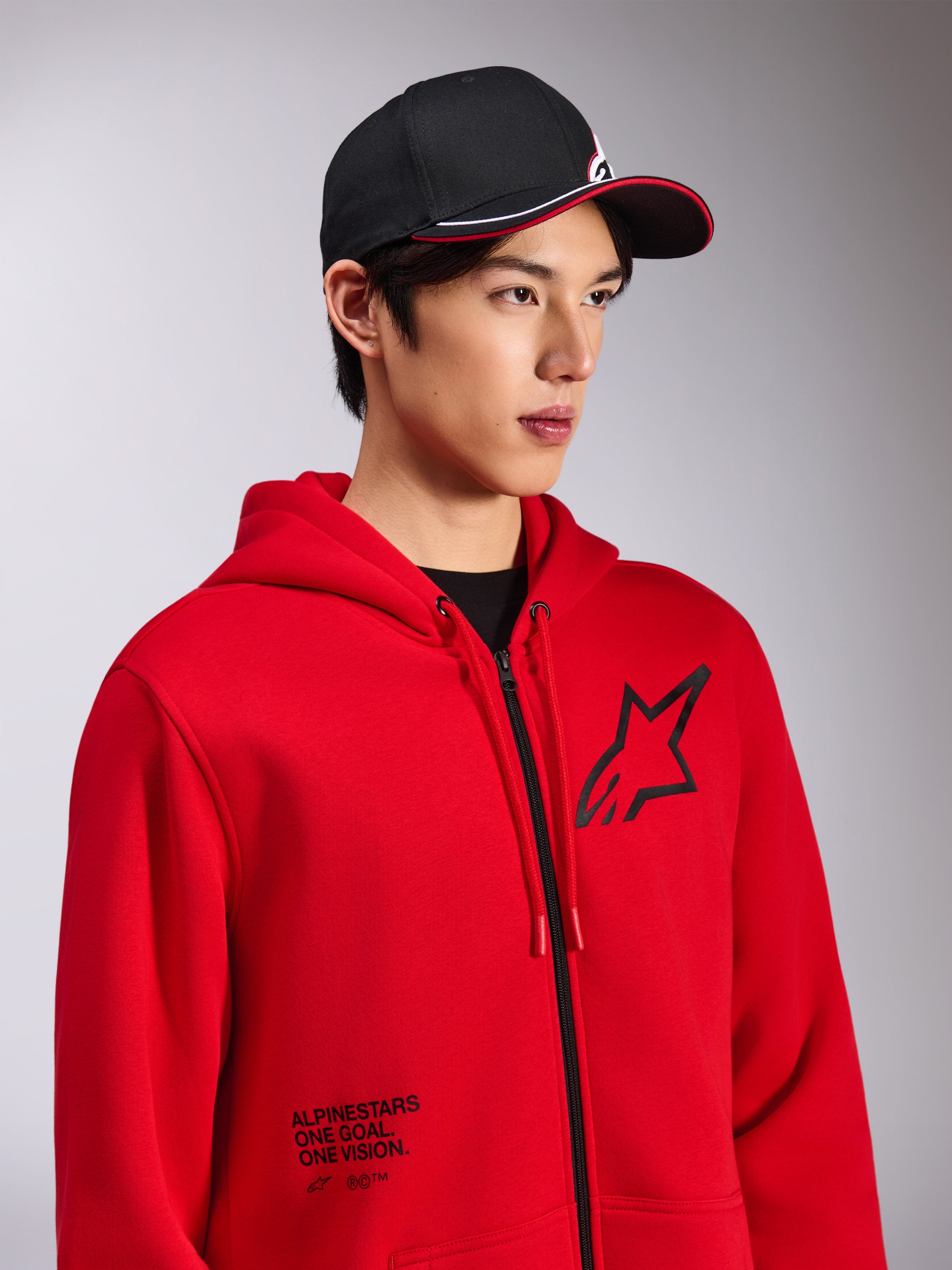 Sweat à capuche Alpinestars Transverse, polaire rouge à fermeture éclair frontale, logo Astars noir sur la poitrine et texte 'One Goal. One Vision.' sur le bas avant, style streetwear décontracté, porté par un mannequin avec une casquette noire