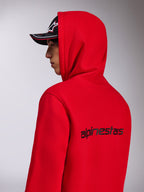 Sweat à capuche Alpinestars Transverse, polaire rouge à capuche, vue de profil arrière montrant le logo Alpinestars noir dans le dos, porté avec la capuche relevée et une casquette noire, style streetwear décontracté inspiré du sport automobile