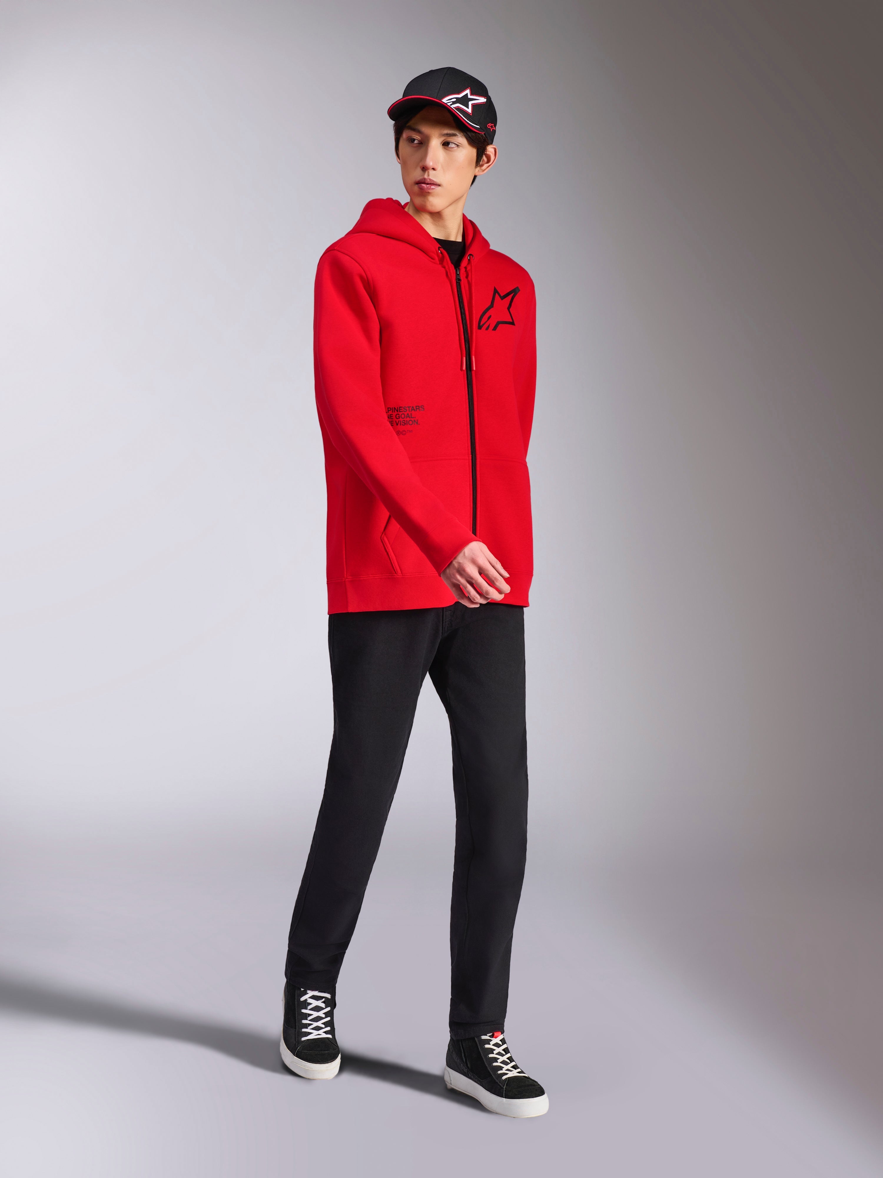Sweat à capuche Alpinestars Transverse rouge, mannequin masculin portant une polaire à capuche à fermeture éclair intégrale, logo Astars noir et texte imprimé, avec pantalon noir et casquette, style streetwear motorsport décontracté