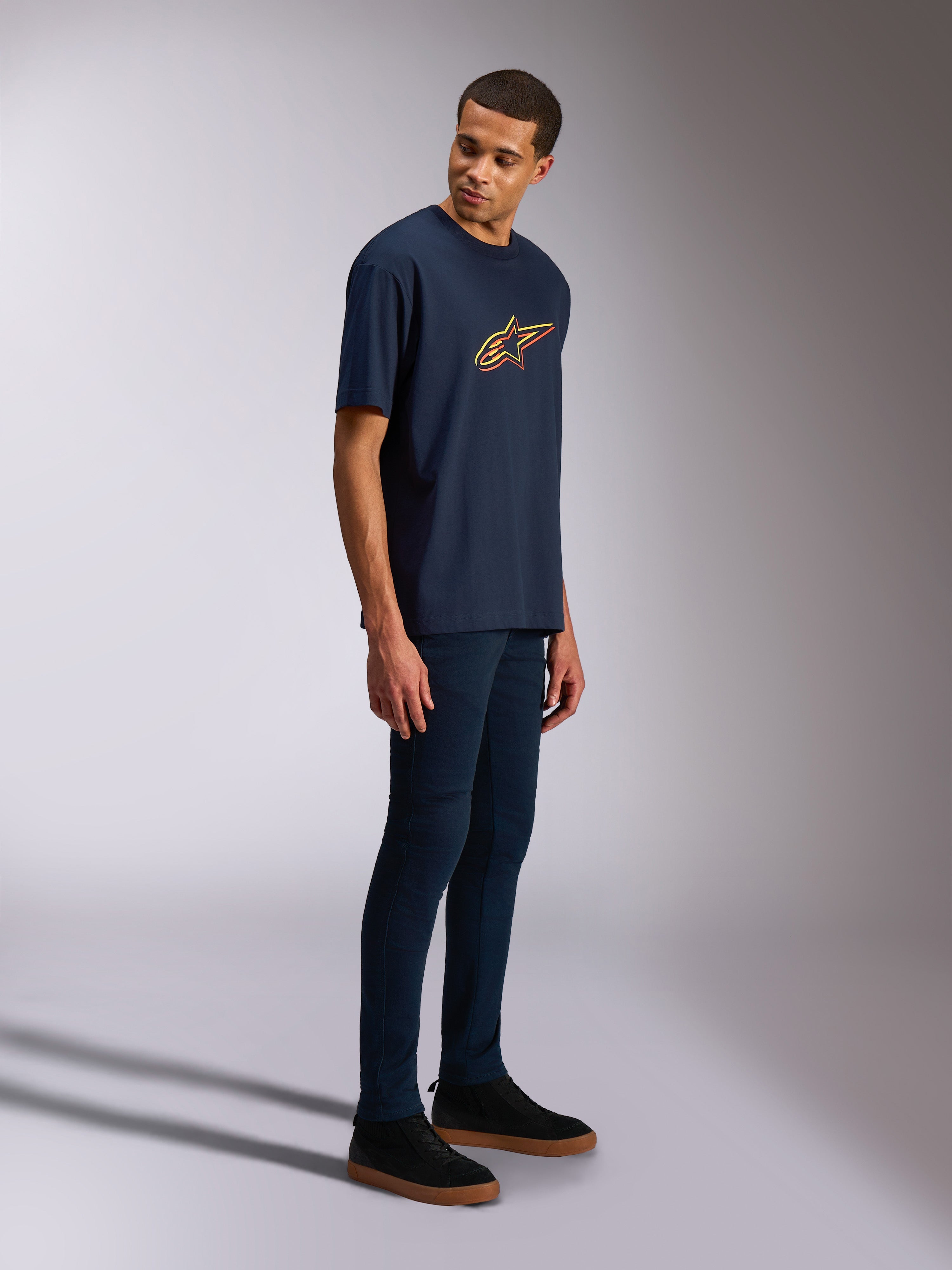 Alpinestars Alter Oversized CSF Tee, T-shirt met korte mouwen, marineblauw, geel en oranje Astars-logo, gedragen door een model met donkere jeans en zwarte sneakers, casual op motorsport geïnspireerde streetwear