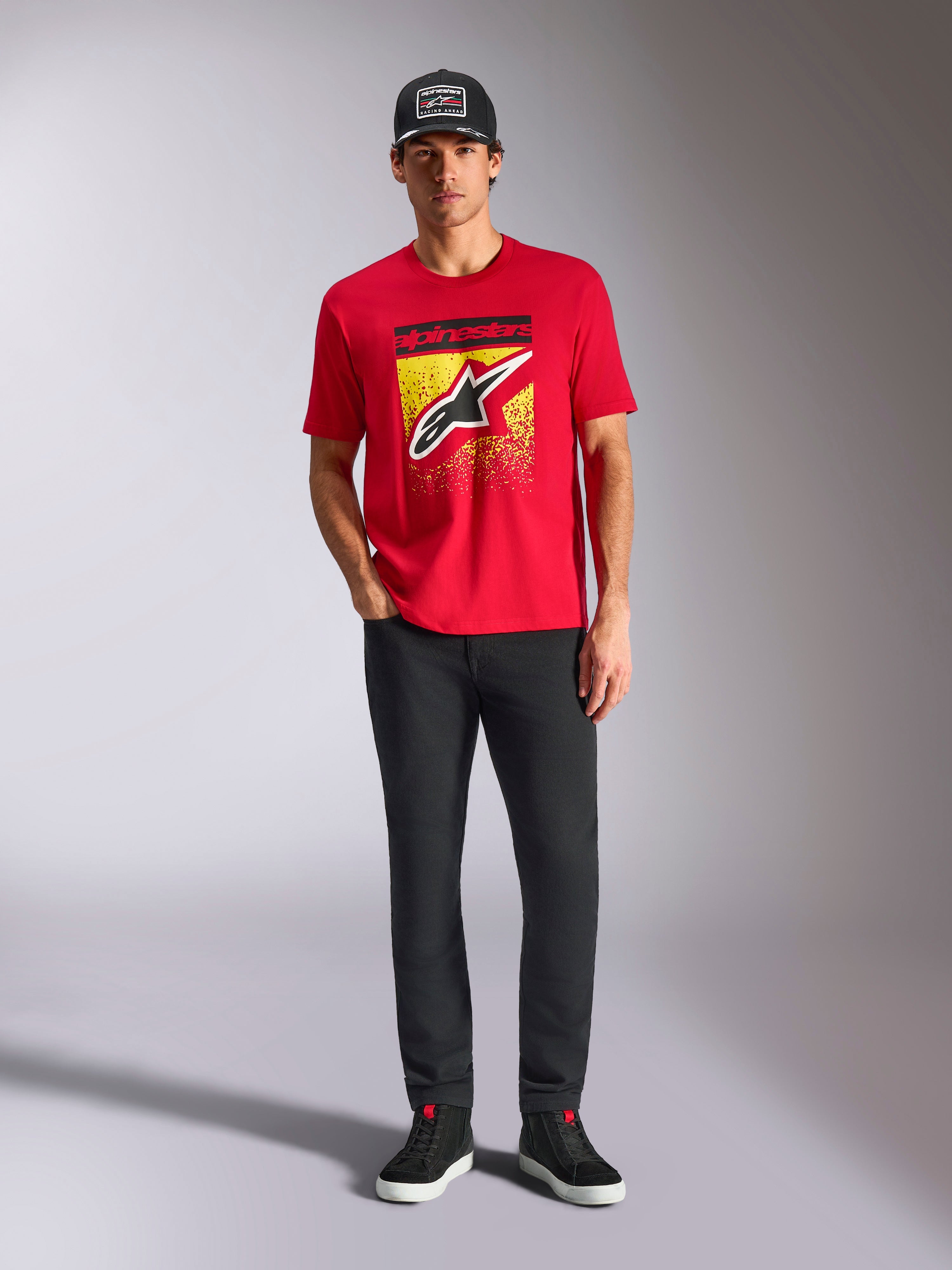 Alpinestars Noka CSF Tee, rood T-shirt met korte mouwen, voorzien van een groot Astars-logo op de voorkant met een geel-zwarte MX-kleurovergang, gedragen met een zwarte broek en zwarte pet, casual motorsport streetwear