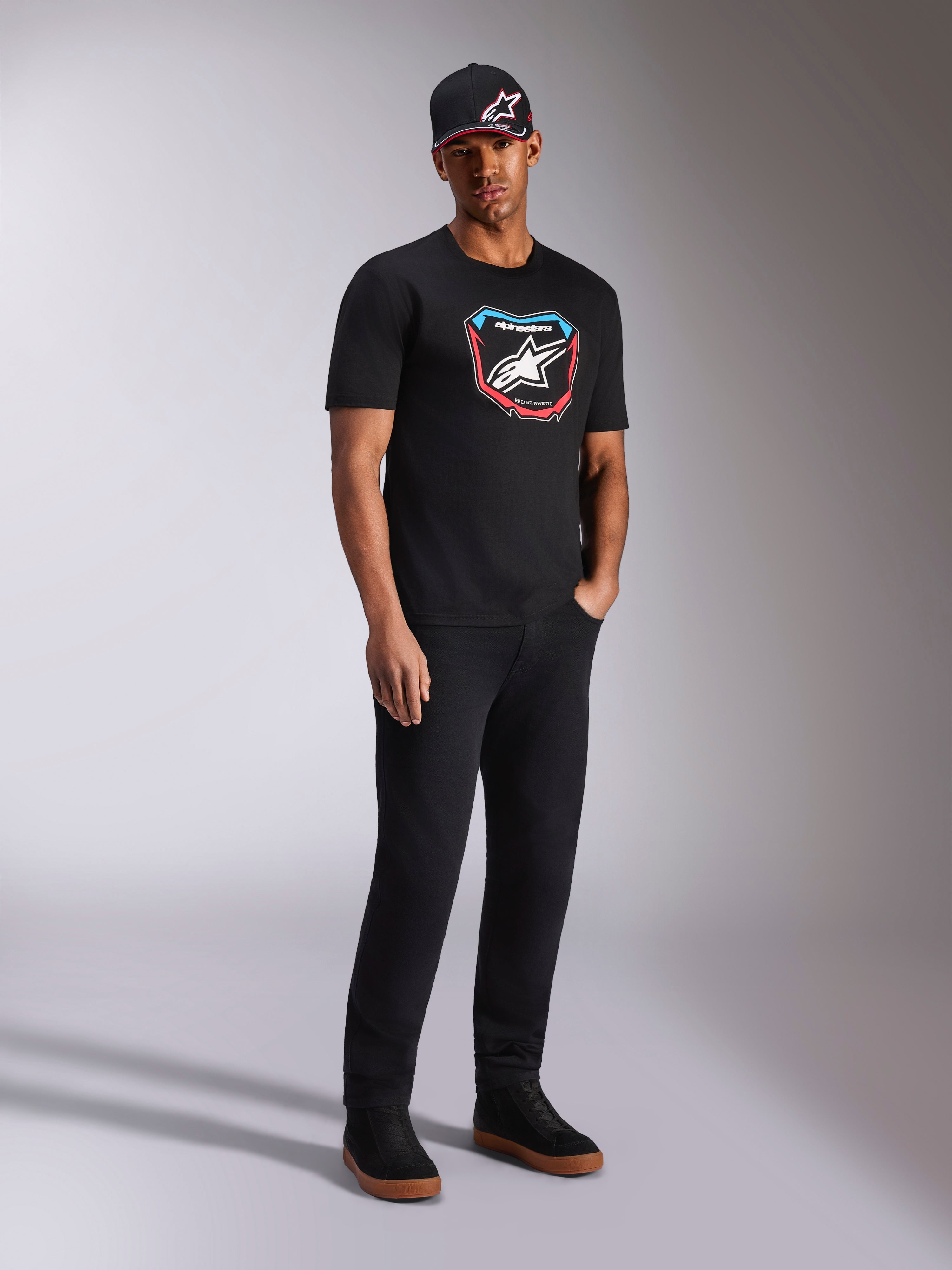 Alpinestars Plate CSF Tee, T-shirt met korte mouwen, zwart, kleurrijke MX-plaat graphic met sterlogo aan de voorzijde, gedragen door model met zwarte logopet en broek, casual motorsport-geïnspireerde streetwear