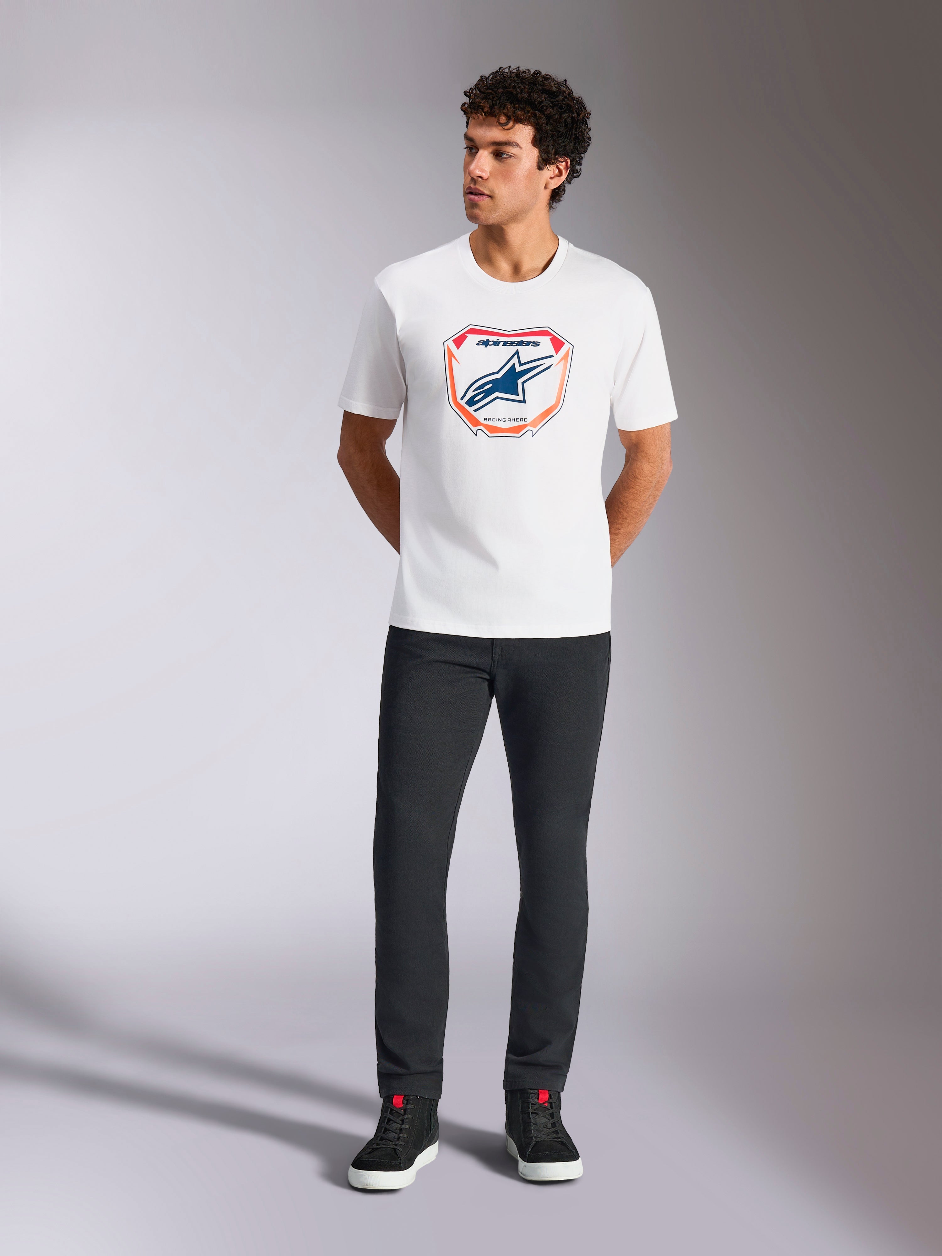 Alpinestars Plate CSF Tee, T-shirt met korte mouwen, wit, met een Astars MX plaatgrafiek aan de voorzijde met oranje en blauwe accenten, gedragen door een model met een zwarte broek en sneakers, casual motorsport streetwear