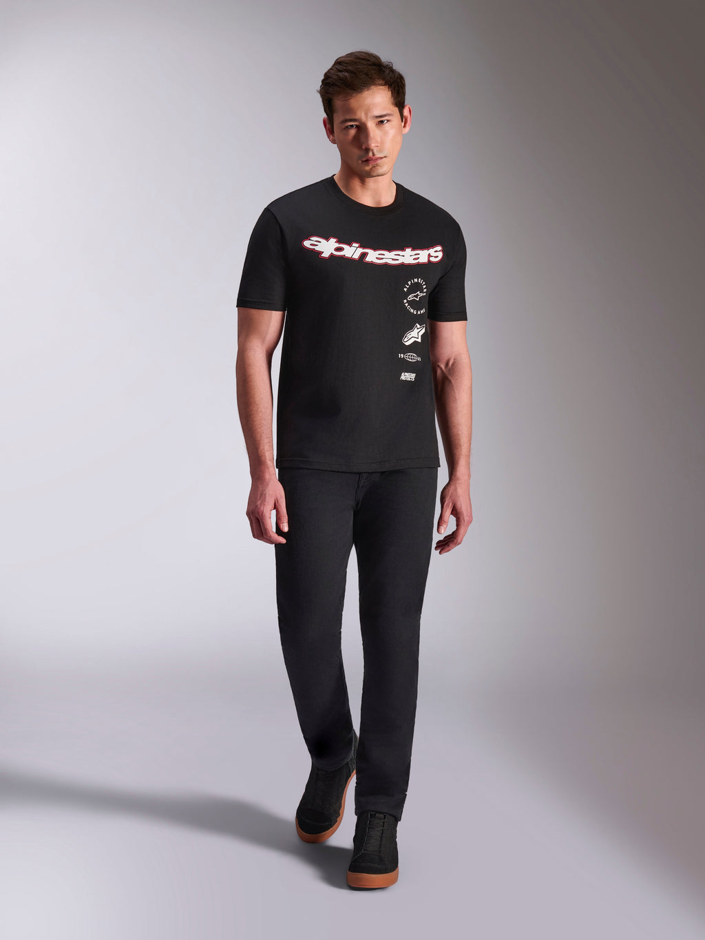 Alpinestars Abus CSF Tee, T-shirt met korte mouwen, zwart, gebogen rood en wit woordmerk met meervoudige gestapelde logo's, gedragen door mannelijk model met zwarte broek en zwarte sneakers, casual motorsport streetwear