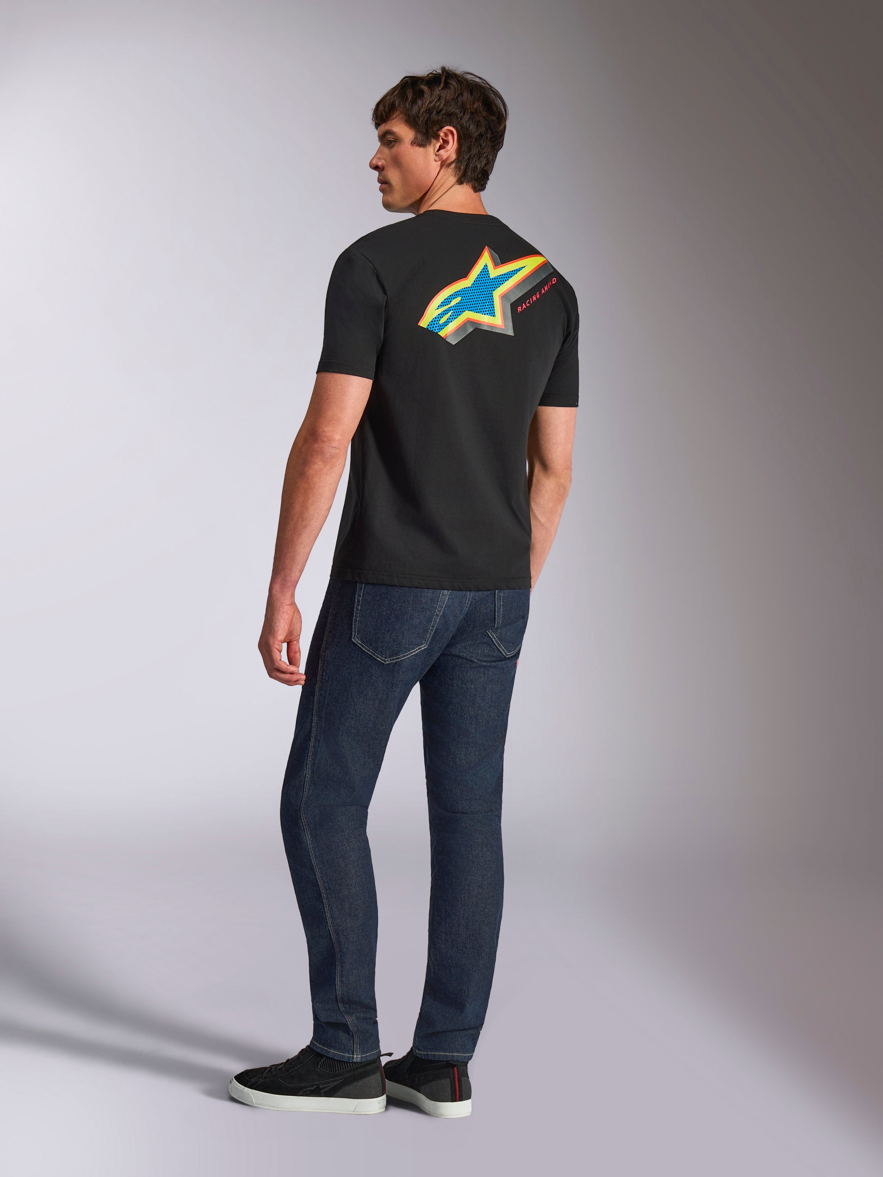 Alpinestars Angler CSF Tee, T-shirt met korte mouwen, zwart, met een groot levendig meerkleurig Astars-logo op de achterkant met blauwe, gele en rode geperforeerde details, gedragen met een donkerblauwe jeans, casual motorsport streetwear