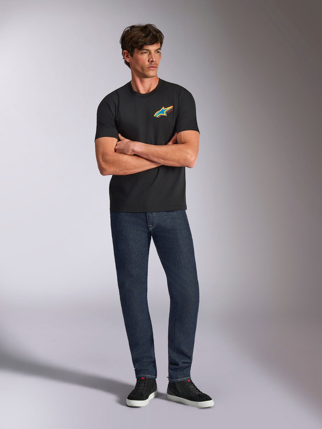 Alpinestars Angler CSF Tee, T-shirt met korte mouwen, zwart, levendig meerkleurig Astars-logo op de borst, gedragen door een model met de armen over elkaar, donkere denim jeans en zwarte sneakers, casual motorsport streetwear