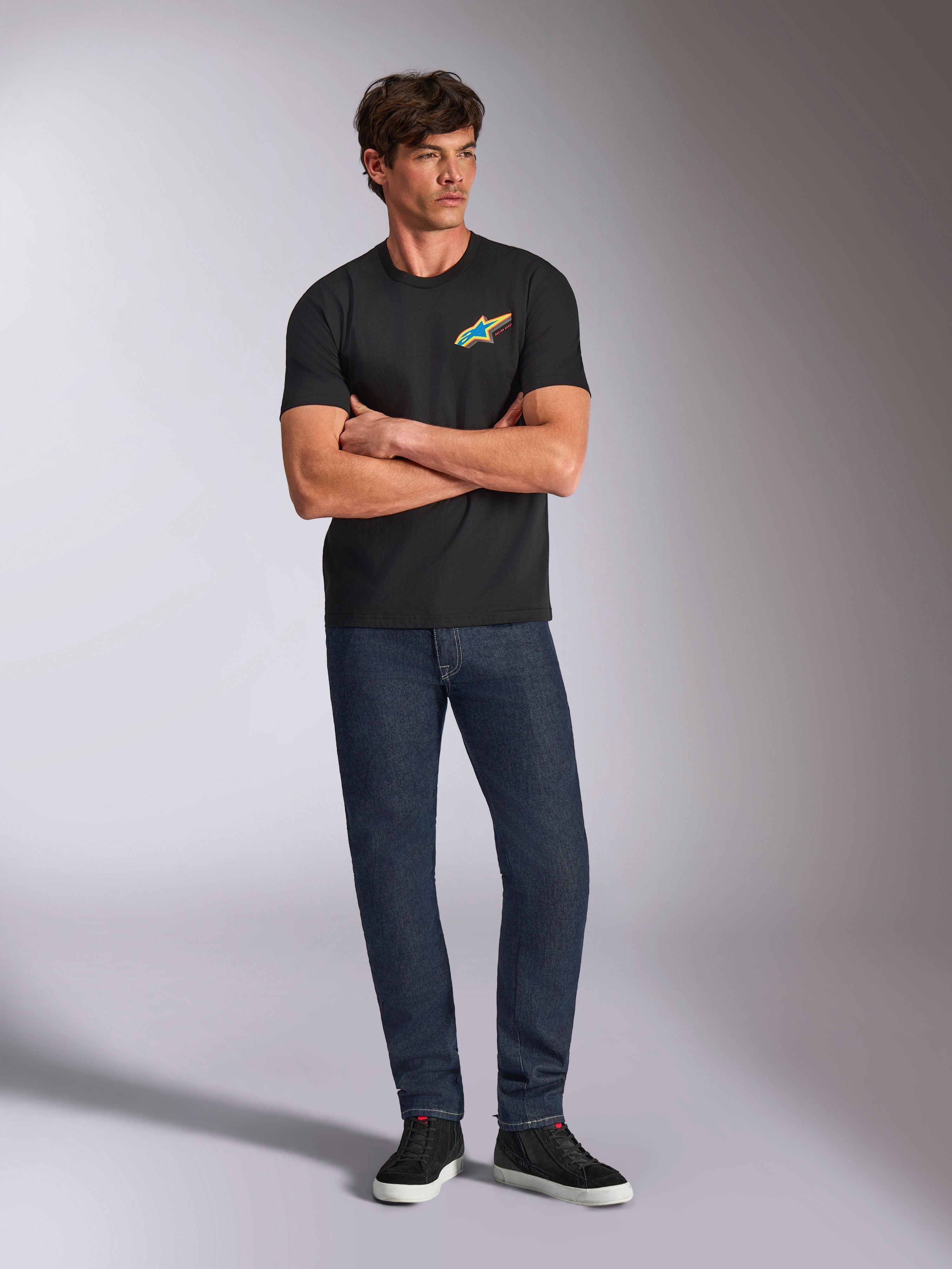 Alpinestars Angler CSF Tee, T-shirt met korte mouwen, zwart, levendig meerkleurig Astars-logo op de borst, gedragen door een model met de armen over elkaar, donkere denim jeans en zwarte sneakers, casual motorsport streetwear