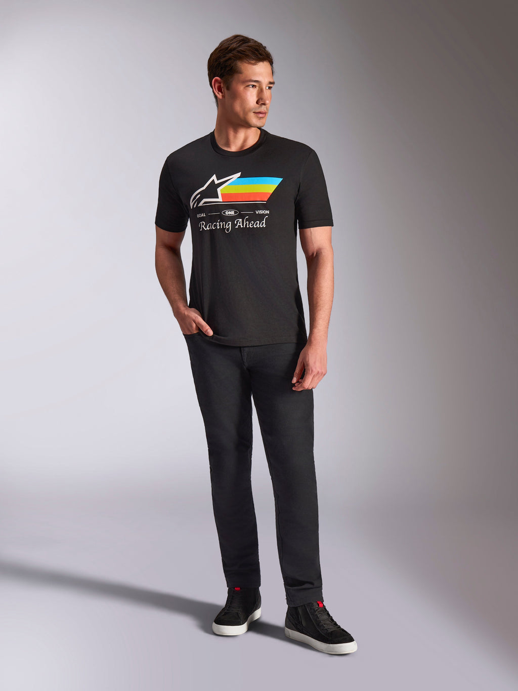 Alpinestars Trifecta CSF Tee, T-shirt met korte mouwen, zwart, met een wit Astars-logo met cyaan, gele en oranje strepen en 'Racing Ahead'-tekst, gedragen door een model met een zwarte broek en sneakers voor casual motorsport streetwear