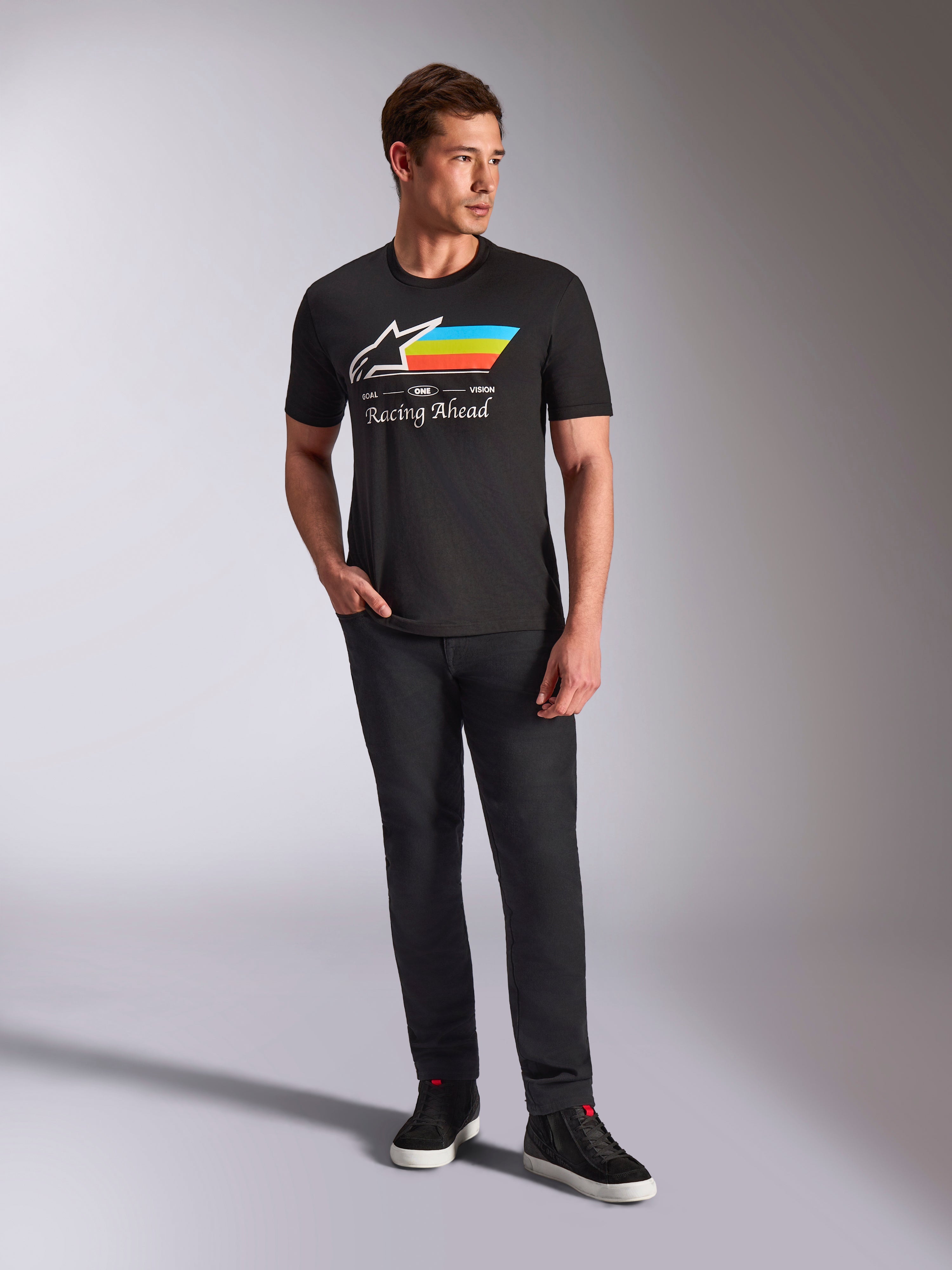 Alpinestars Trifecta CSF Tee, T-shirt met korte mouwen, zwart, met een wit Astars-logo met cyaan, gele en oranje strepen en 'Racing Ahead'-tekst, gedragen door een model met een zwarte broek en sneakers voor casual motorsport streetwear