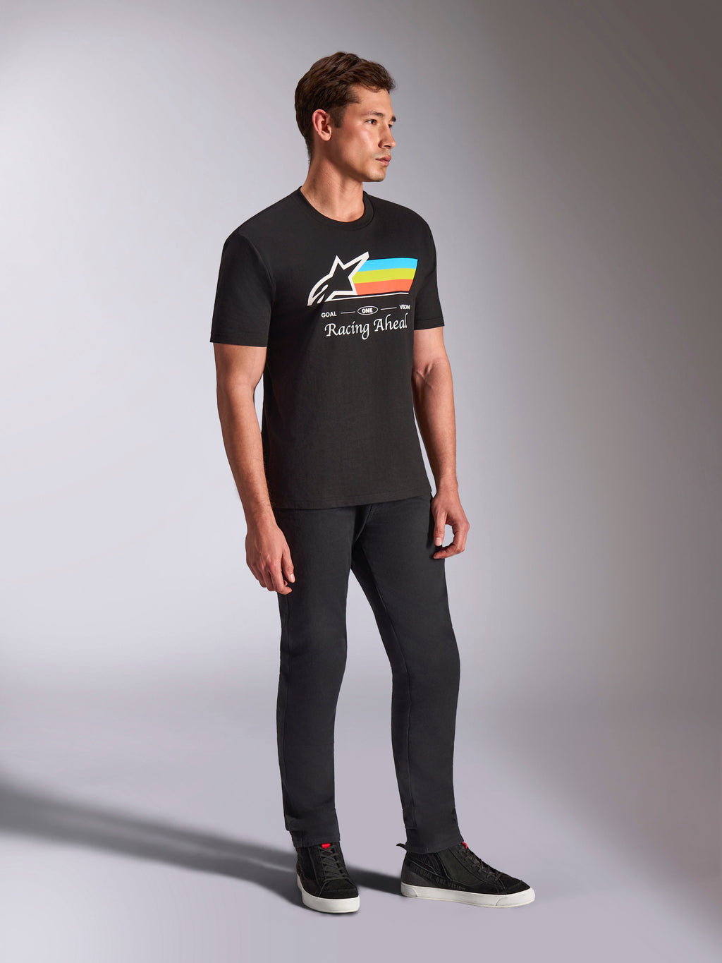 Alpinestars Trifecta CSF Tee, T-shirt met korte mouwen, zwart, wit Astars-logo met blauwe, gele en oranje strepen en 'Racing Ahead'-graphic, gedragen door een model met een zwarte broek en sneakers, casual motorsport-geïnspireerde lifestyle-kleding