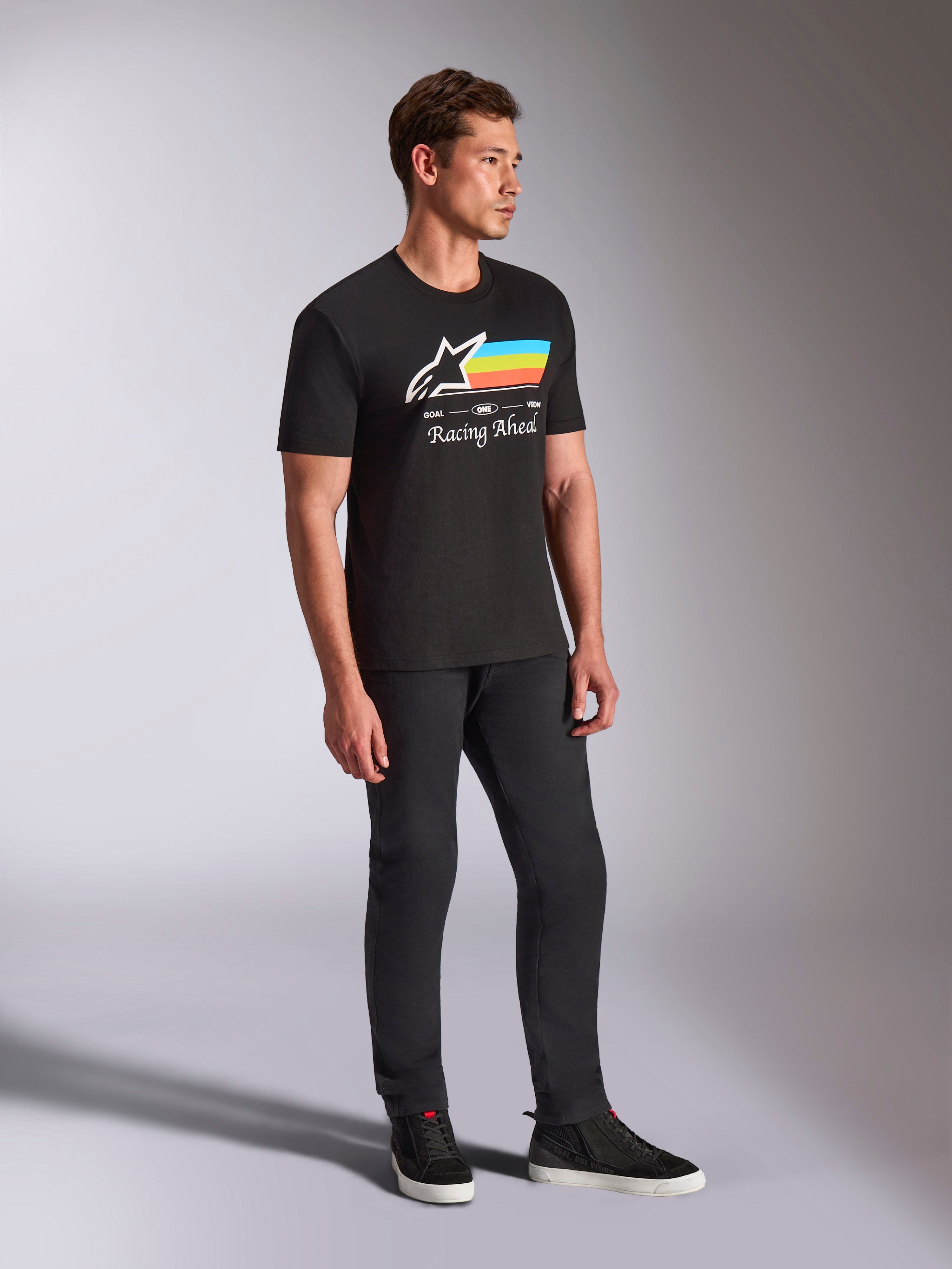Alpinestars Trifecta CSF Tee, T-shirt met korte mouwen, zwart, wit Astars-logo met blauwe, gele en oranje strepen en 'Racing Ahead'-graphic, gedragen door een model met een zwarte broek en sneakers, casual motorsport-geïnspireerde lifestyle-kleding