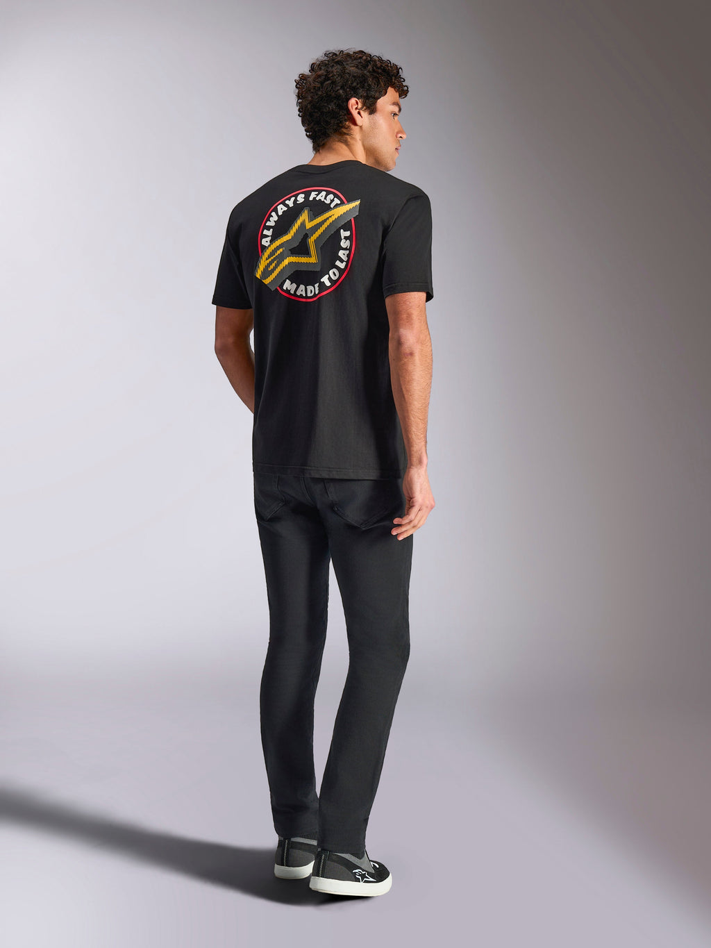 Alpinestars Aorbit CSF Tee, T-shirt met korte mouwen, zwart, achteraanzicht met een groot geel Astars-logo en ronde 'Always Fast Made to Last' grafische print, gedragen met zwarte jeans en casual sneakers, motorsport-geïnspireerde streetwear-stijl