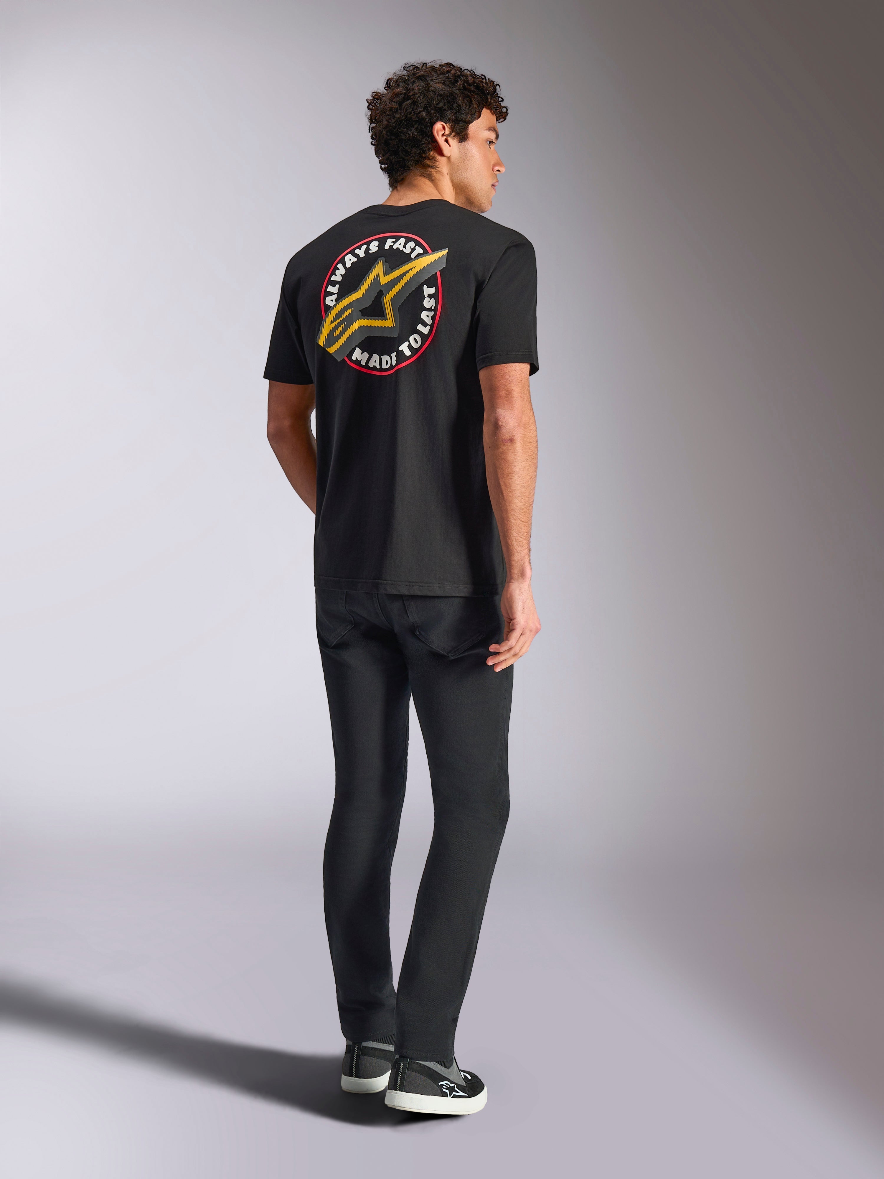 Alpinestars Aorbit CSF Tee, T-shirt met korte mouwen, zwart, achteraanzicht met een groot geel Astars-logo en ronde 'Always Fast Made to Last' grafische print, gedragen met zwarte jeans en casual sneakers, motorsport-geïnspireerde streetwear-stijl