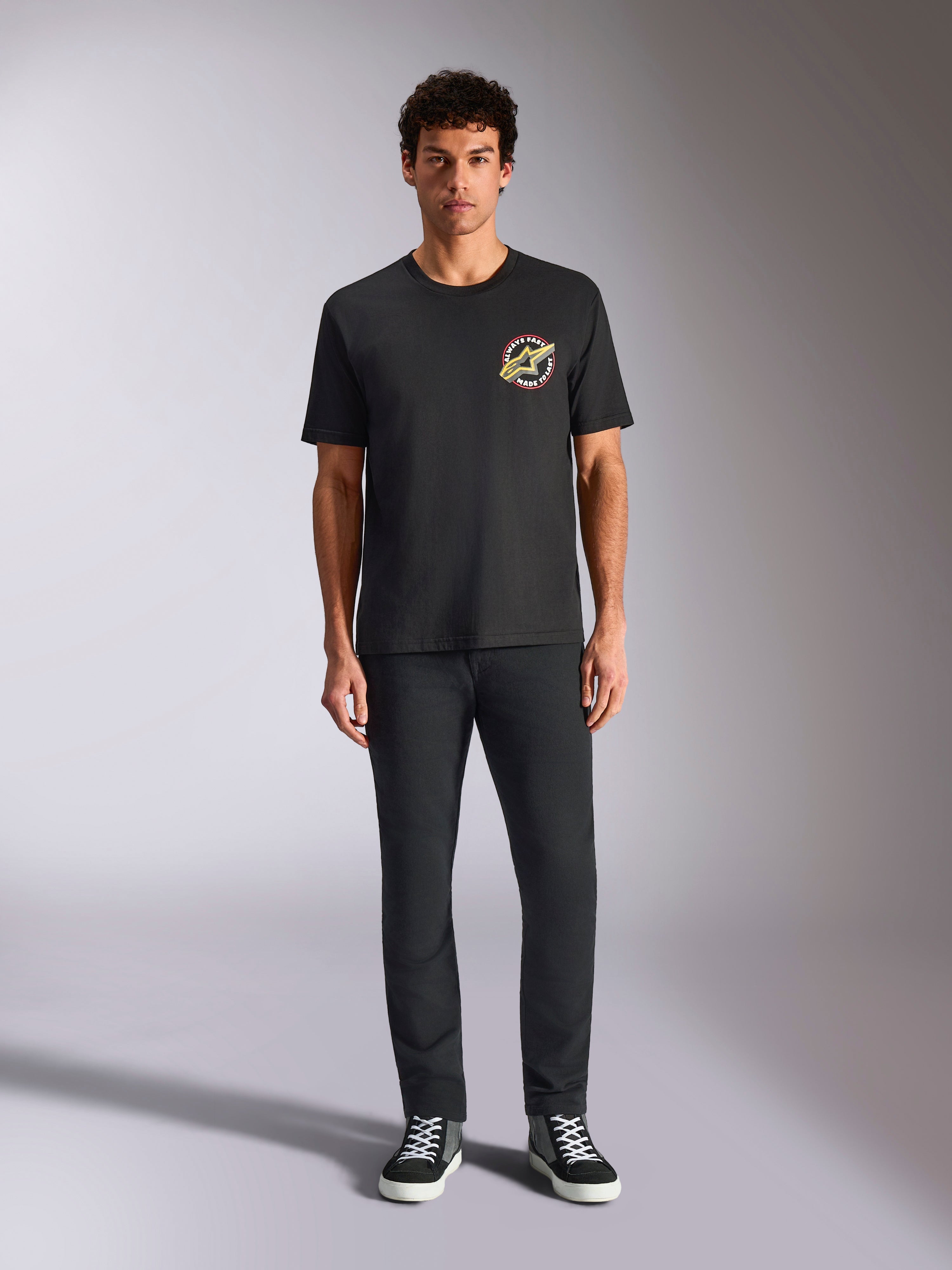 Alpinestars Aorbit CSF Tee, T-shirt met korte mouwen, zwart, circulair logo met 'Always Fast Made to Last'-tekst op de borst, gedragen door een model met zwarte broek en sneakers, casual motorsport-geïnspireerde streetwear