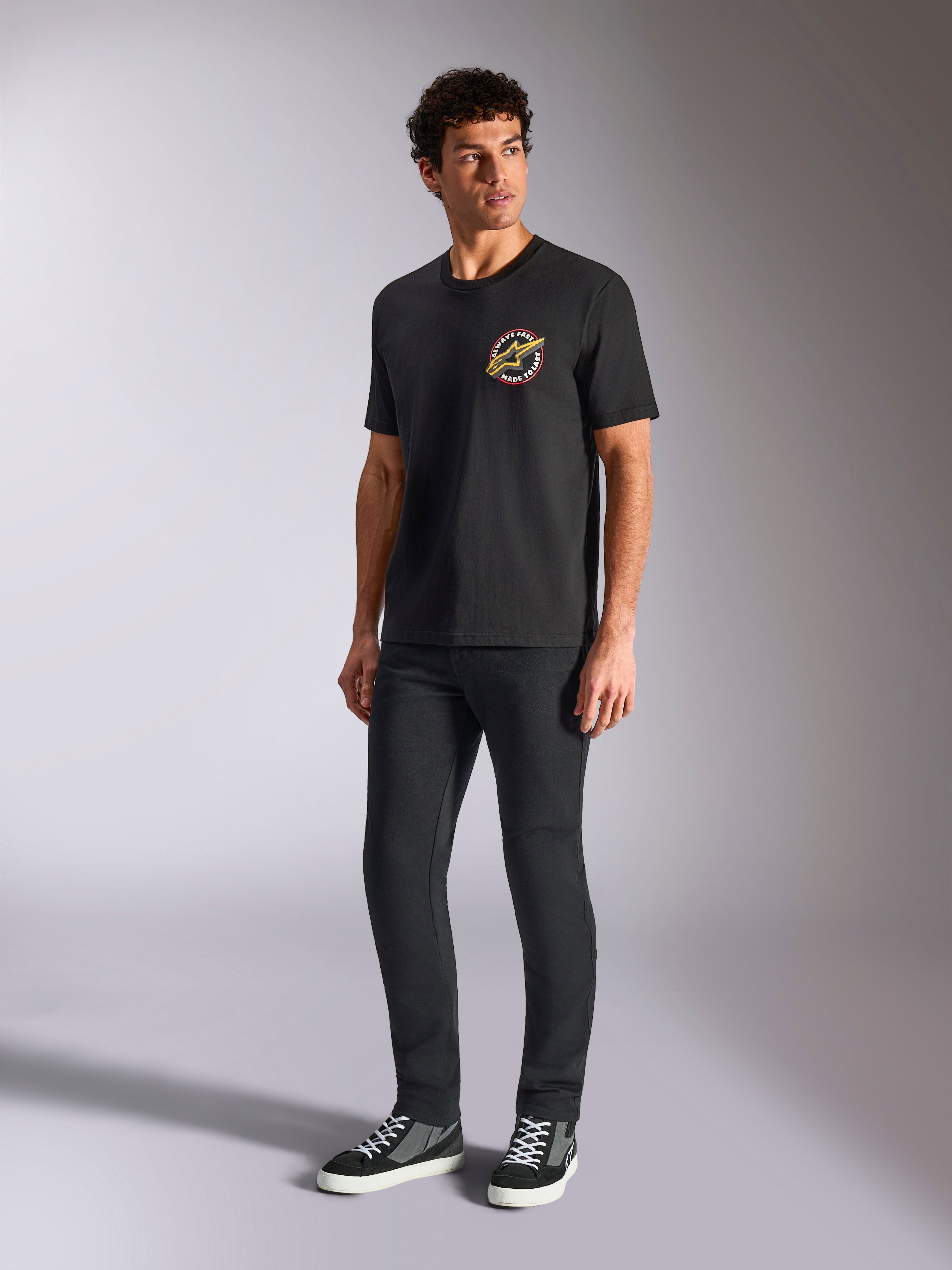 Alpinestars Aorbit CSF Tee, t-shirt met korte mouwen, zwart, met een ronde Always Fast Made to Last-graphic op de borst, gedragen door een model met een donkere slim-fit broek en sneakers, casual motorsport streetwear