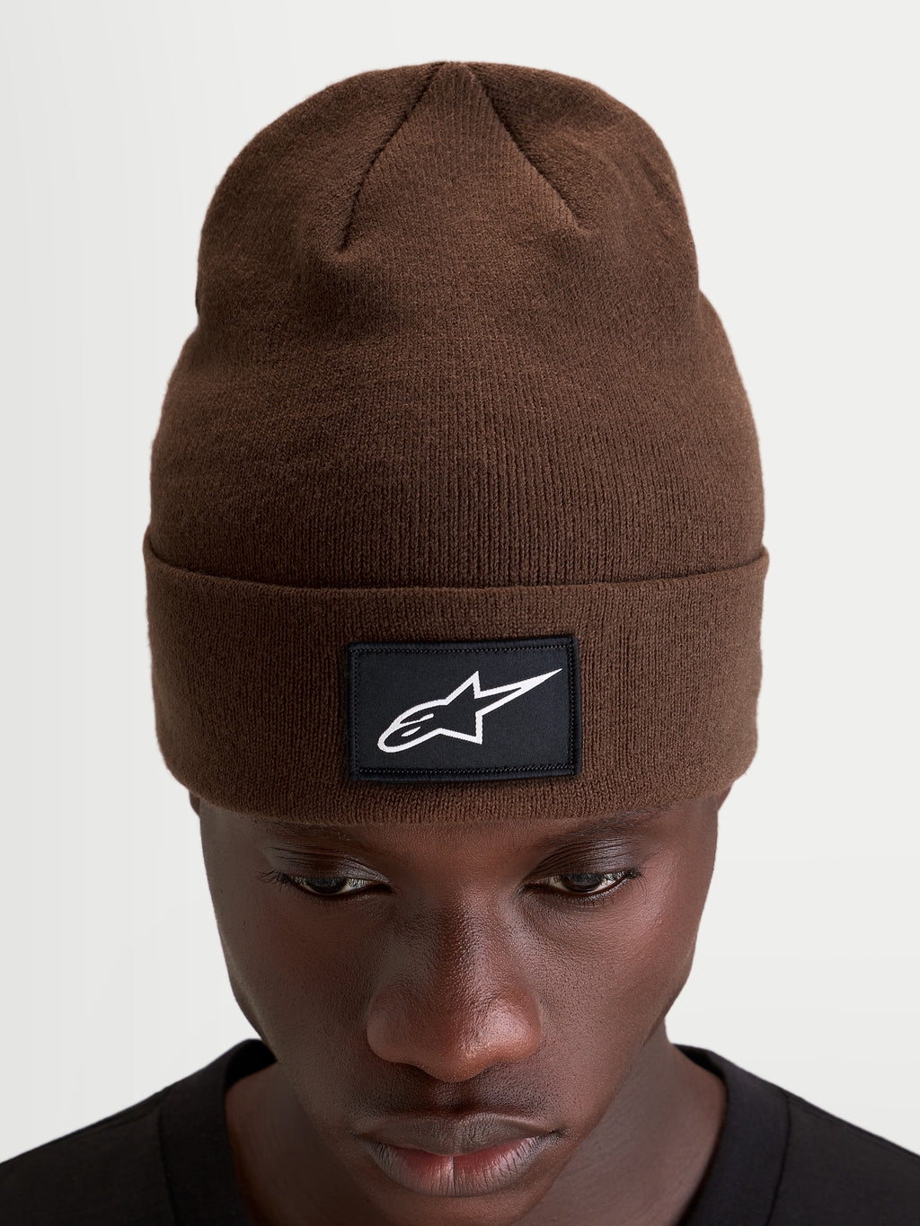 Alpinestars Augment Cuff Beanie, knit sportswear winter hat in brown, featuring a woven black patch with a white Astars logo and a deep-fit pull-on silhouette worn on a model
