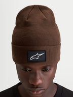 Alpinestars Augment Cuff Beanie, knit sportswear winter hat in brown, featuring a woven black patch with a white Astars logo and a deep-fit pull-on silhouette worn on a model