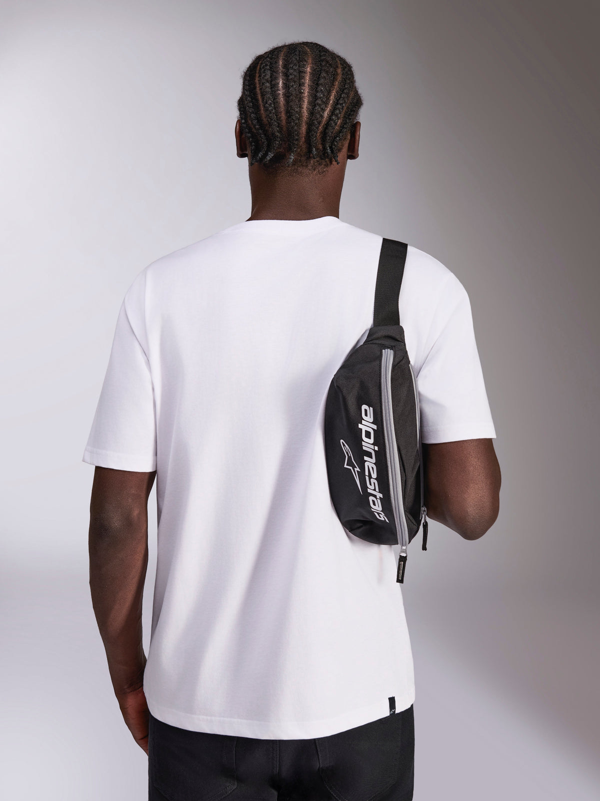 Alpinestars Only Linear heuptas, zwart met wit Alpinestars-logo, gedragen als crossbodytas over een wit T-shirt, casual motorsport-geïnspireerd streetwear-accessoire