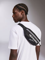 Sacoche de hanche Alpinestars Only Linear, noir et blanc, portée en bandoulière sur un t-shirt blanc, avec logo Alpinestars blanc et fermeture éclair verticale, accessoire streetwear compact.