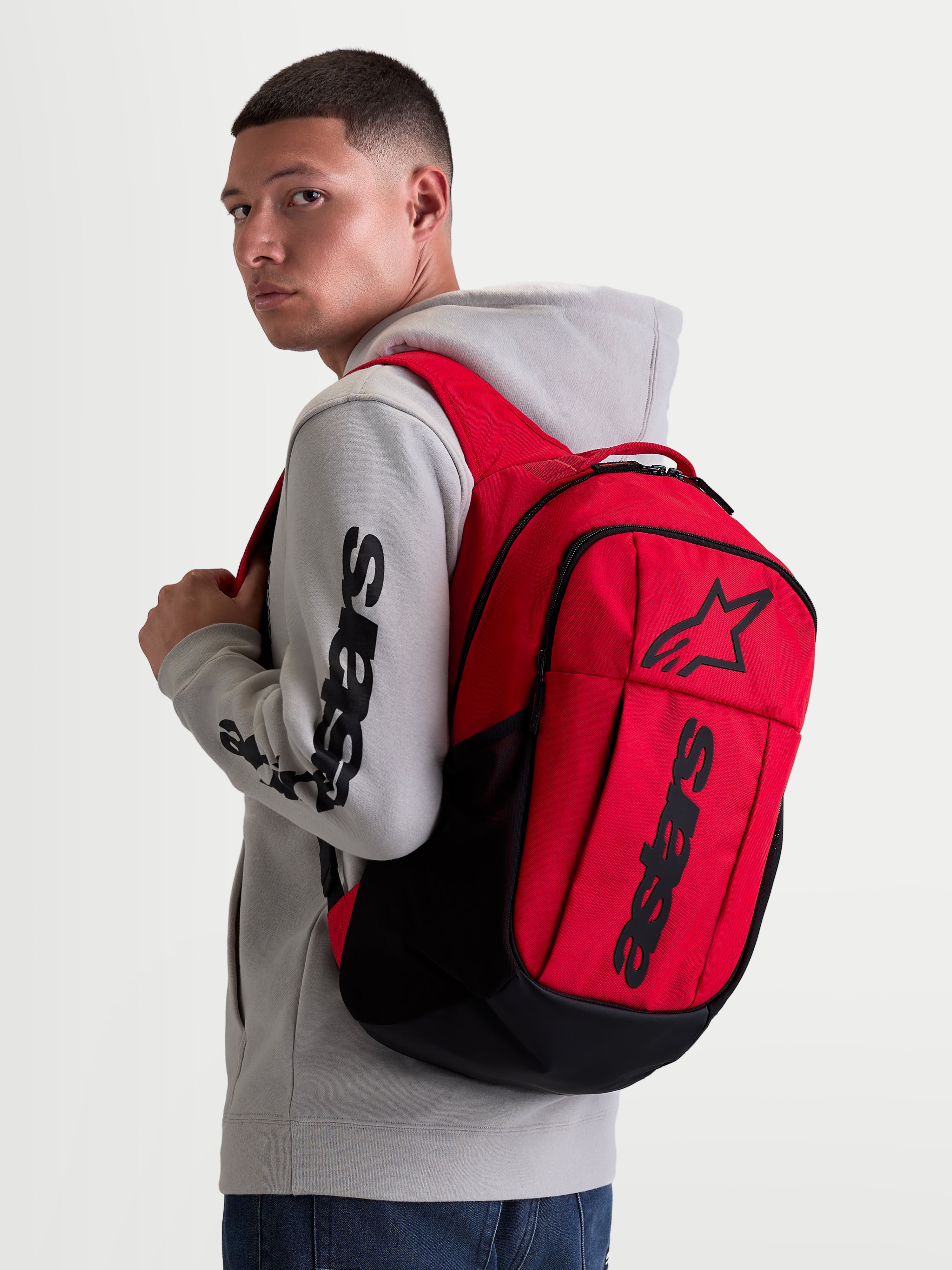Alpinestars GFX Boss Rucksack, 20L Urban Sportswear Tagesrucksack, Rot und Schwarz, am Modell in grauem Kapuzenpullover gezeigt. Mit strapazierfähiger 600D Polyester-Konstruktion, erhabenem Silikon-Astars-Grafik-Branding und internem Laptopfach. Ideal für das tägliche Pendeln und den Alltag.