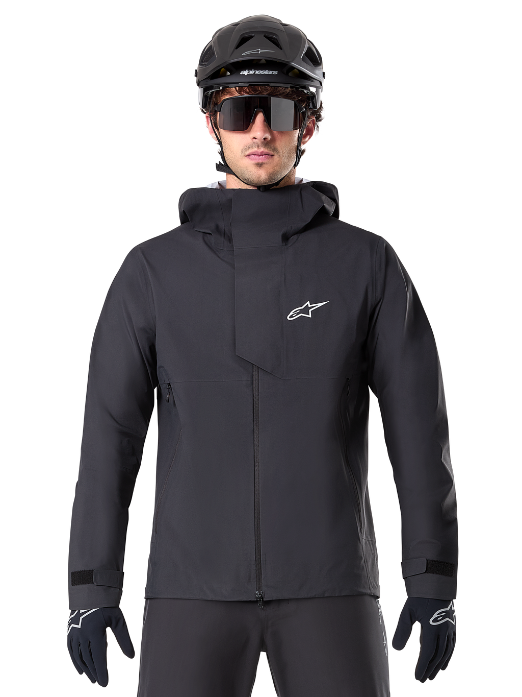 Veste imperméable Alpinestars A-Dura Elite, noire, veste de VTT, design à col montant avec capuche, fermeture éclair frontale avec rabat de protection et logo blanc, portée avec un casque noir et des gants de cyclisme, protection technique pour le trail