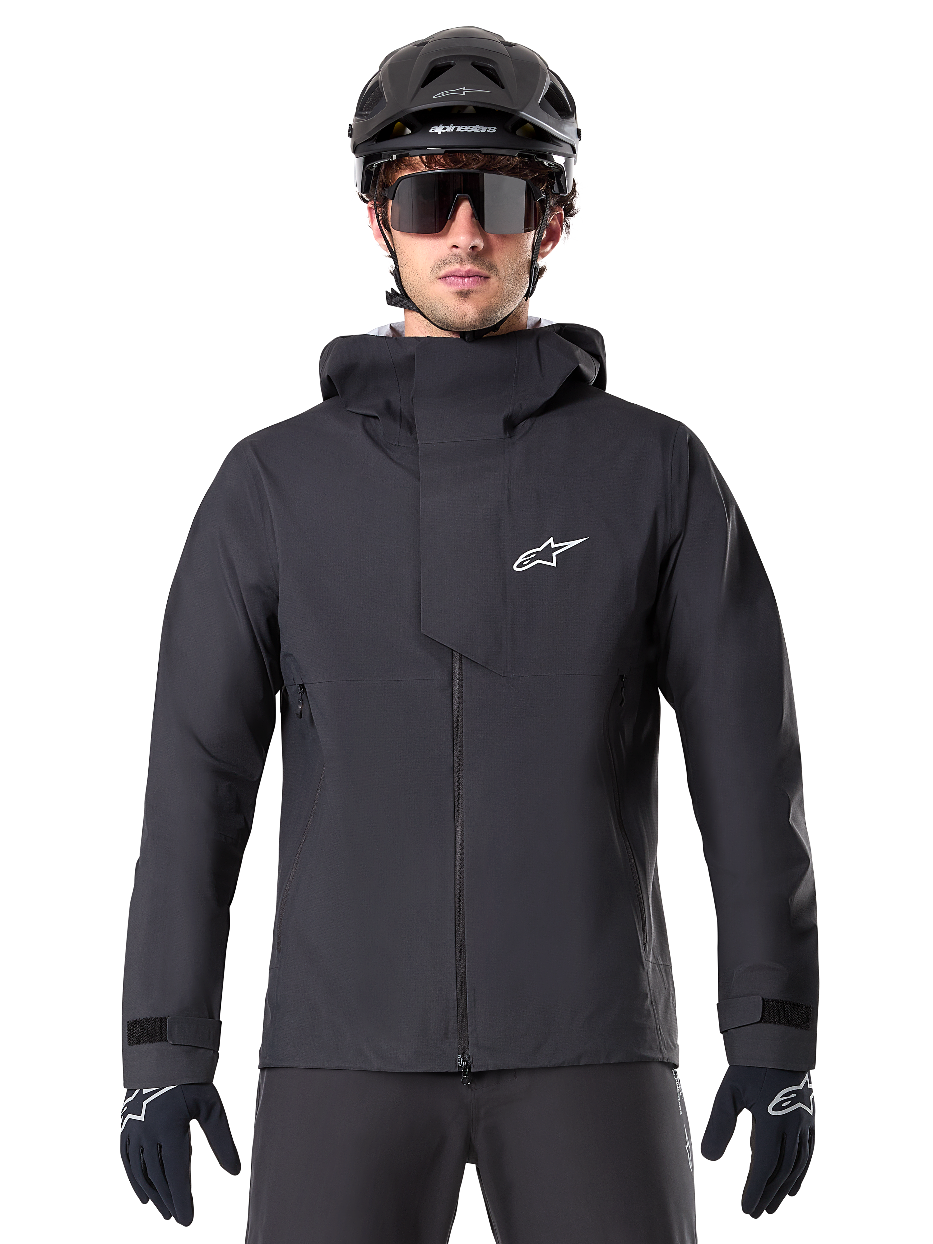 Veste imperméable Alpinestars A-Dura Elite, noire, veste de VTT, design à col montant avec capuche, fermeture éclair frontale avec rabat de protection et logo blanc, portée avec un casque noir et des gants de cyclisme, protection technique pour le trail