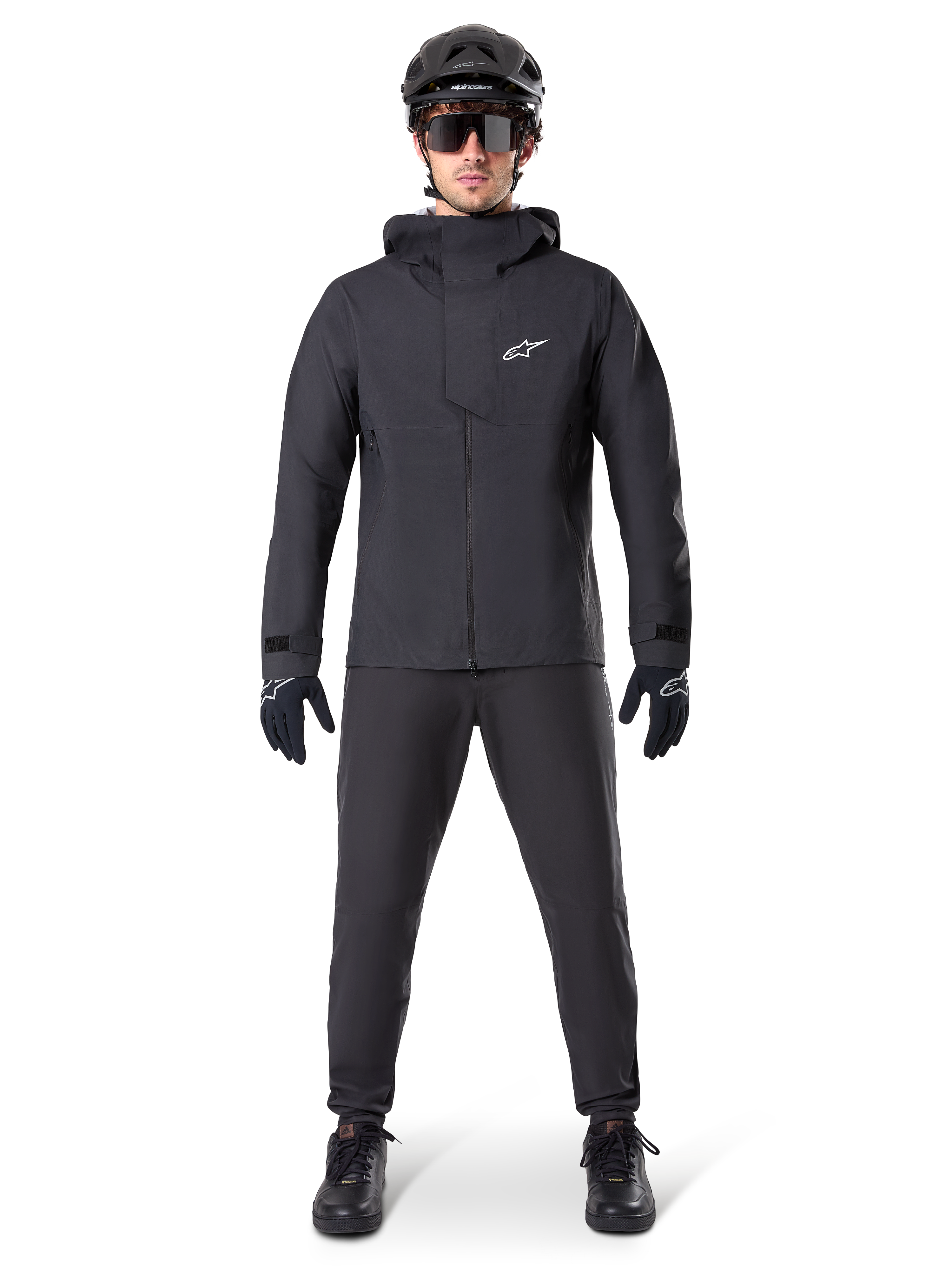Veste imperméable Alpinestars A-Dura Elite, noire, portée avec un pantalon et des gants de cyclisme assortis, casque noir et lunettes de soleil, logo Astars blanc sur la poitrine, design technique haute performance pour le VTT