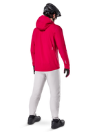 Veste imperméable Alpinestars A-Dura Elite, rouge fluo, vue de dos de trois-quarts, portée avec un pantalon gris clair et un casque de VTT noir, dotée d'une capuche et de détails réfléchissants, veste de pluie technique haute performance pour le trail