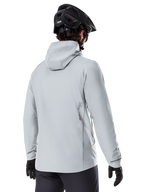 Alpinestars A-Dura Thermal Jacket, Titanium grijs, achteraanzicht van een rijder die de geïsoleerde jas met capuchon draagt, gecombineerd met een zwarte MTB-helm en zwarte handschoenen, met een klein Alpinestars-logo op de onderrug, ontworpen voor mountainbiken in koud weer