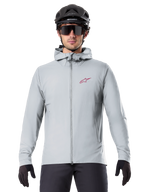 Alpinestars A-Dura Thermal Jacket in Titanium, lichtgrijze fietsjas met capuchon, rood sterlogo op de borst, gedragen door een rijder met zwarte MTB-helm, zonnebril en handschoenen, geïsoleerde kleding voor mountainbiken